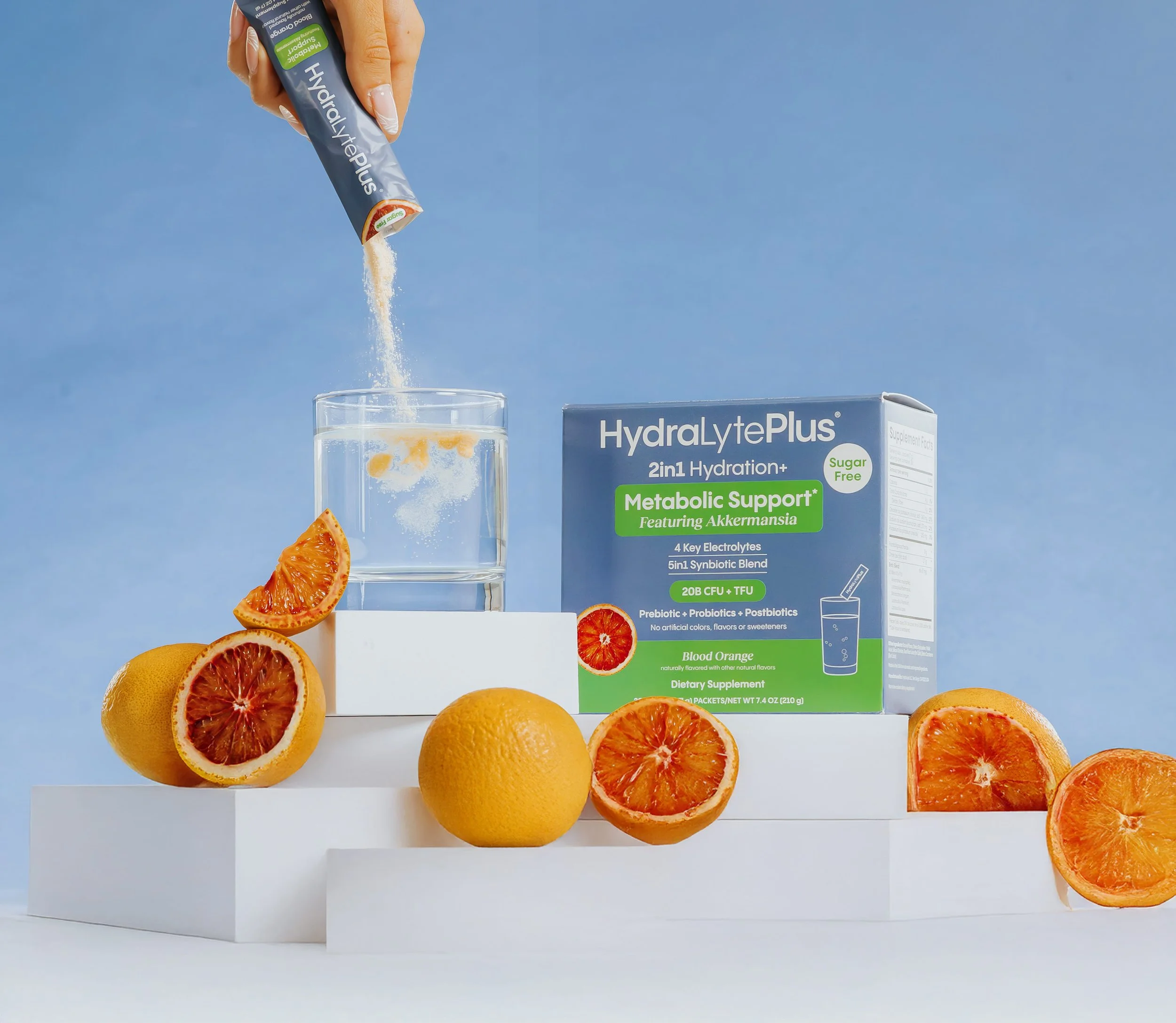 Several fresh blood oranges, some halved and some whole, are arranged on a white display. A box of HydralytePlus supplement with orange flavor is positioned behind the oranges. A hand is pouring powder from a packet of HydralytePlus into a clear glass of water, and there are more oranges and slices around the display against a blue background.
