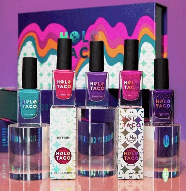 Five bottles of HOLO TACO nail polish in various colors with their boxes behind them, arranged on top of clear display blocks against a colorful background.