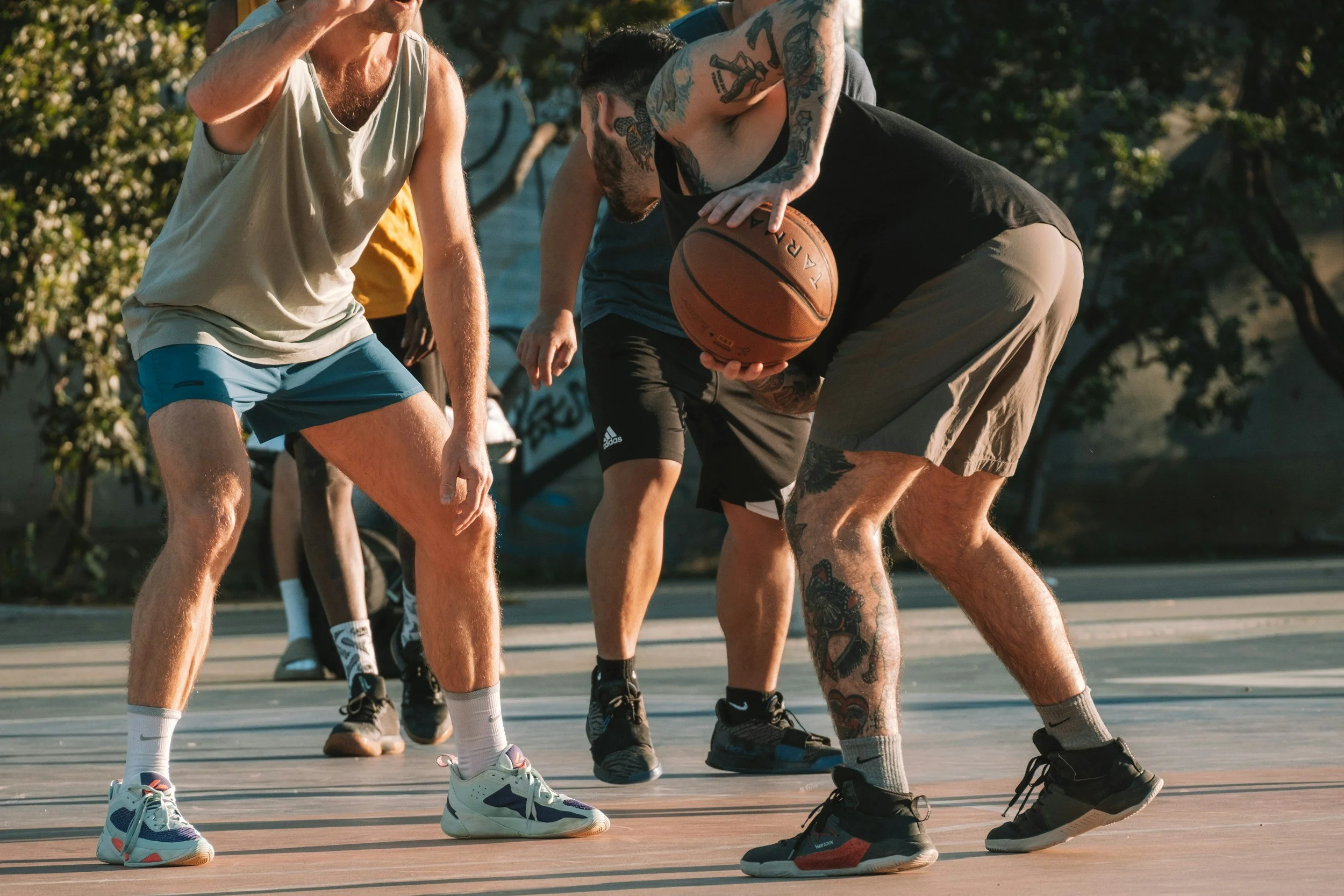 People playing basketball on an outdoor court, with one person dribbling the ball while others defend.