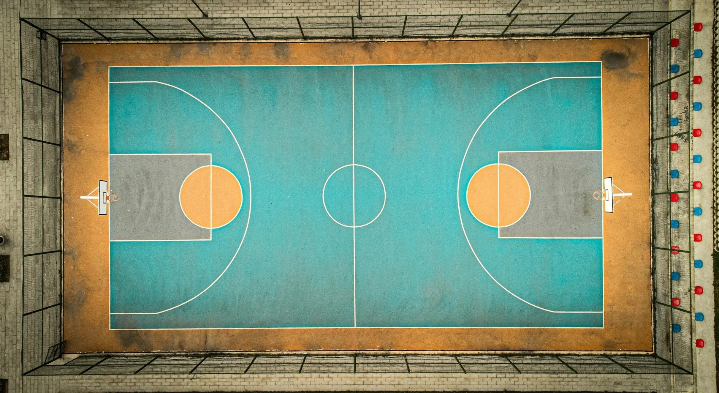 An aerial view of a fenced outdoor basketball court with blue, orange, and gray markings, surrounded by a brick wall and red and blue light posts.