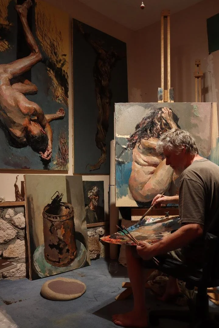 An artist is painting a portrait of himself in a studio filled with completed paintings of nude figures and still life on the walls and floor.