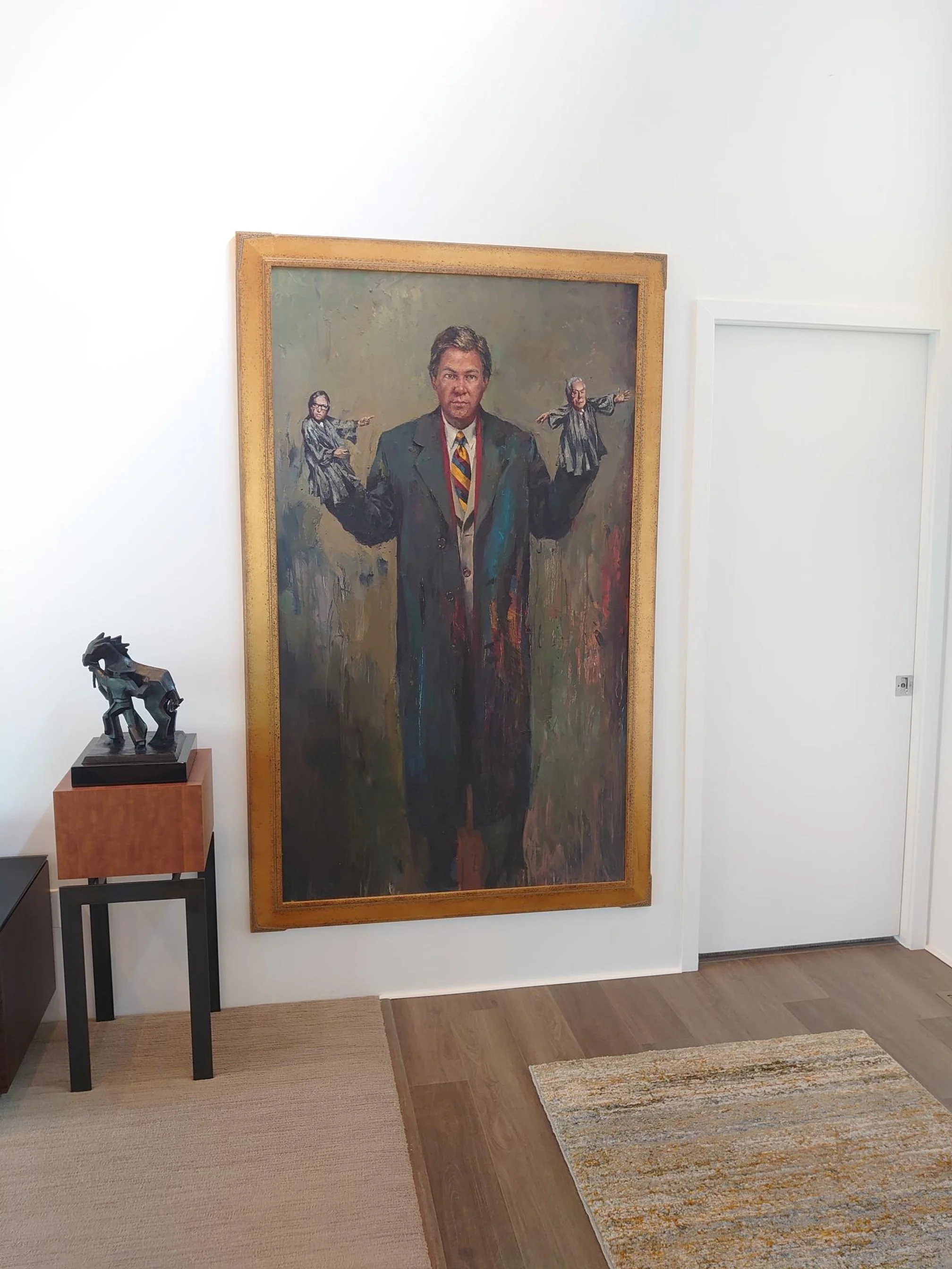 A large portrait painting of a man in a suit and tie, with two smaller figures in robes and glasses painted on either side of him, hung on a white wall in a room with wooden flooring and a small table with a sculpture of a goat or ram.