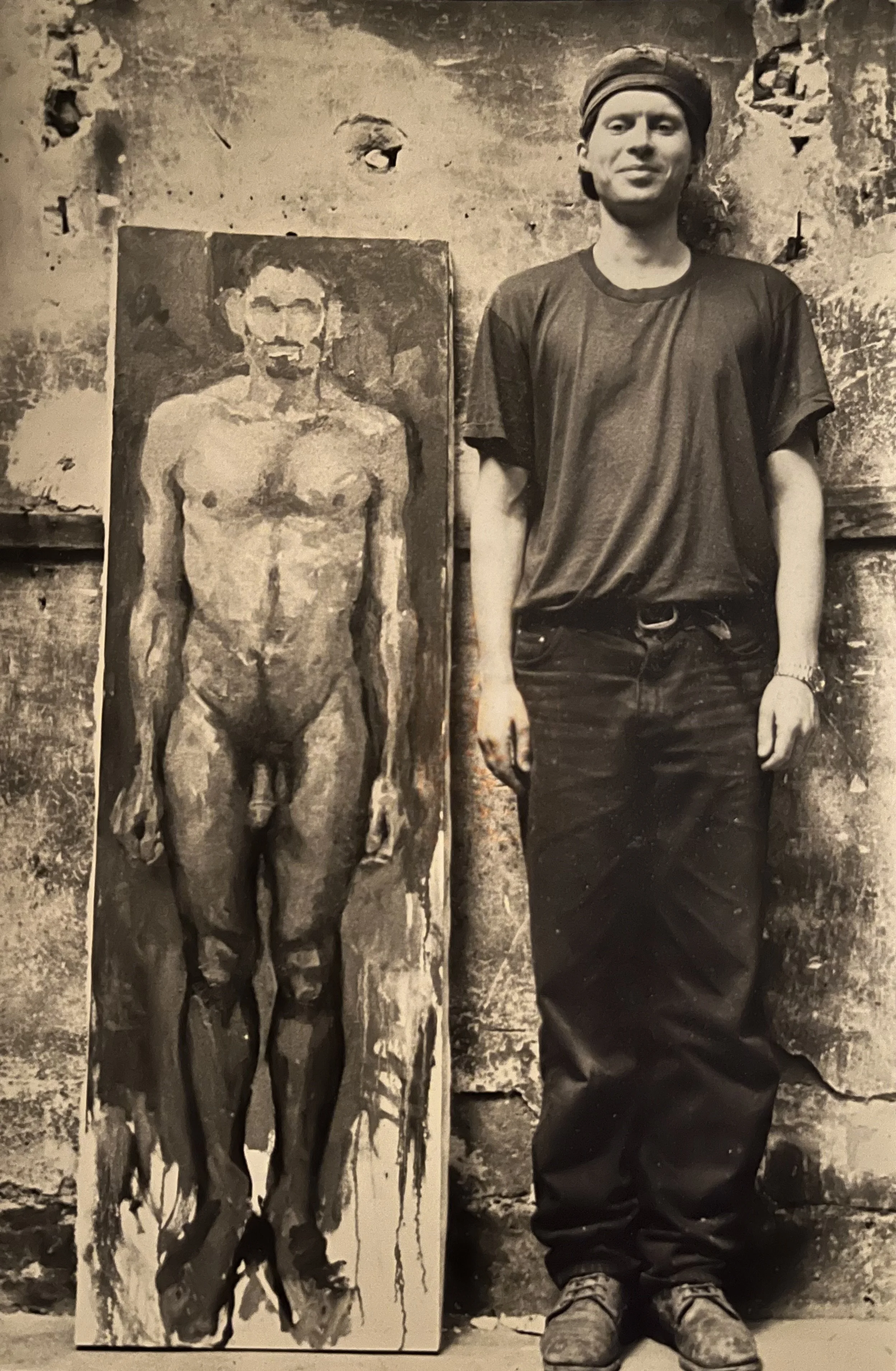 A young man in casual clothing standing next to a painting of a nude male figure on a rough, textured wall.
