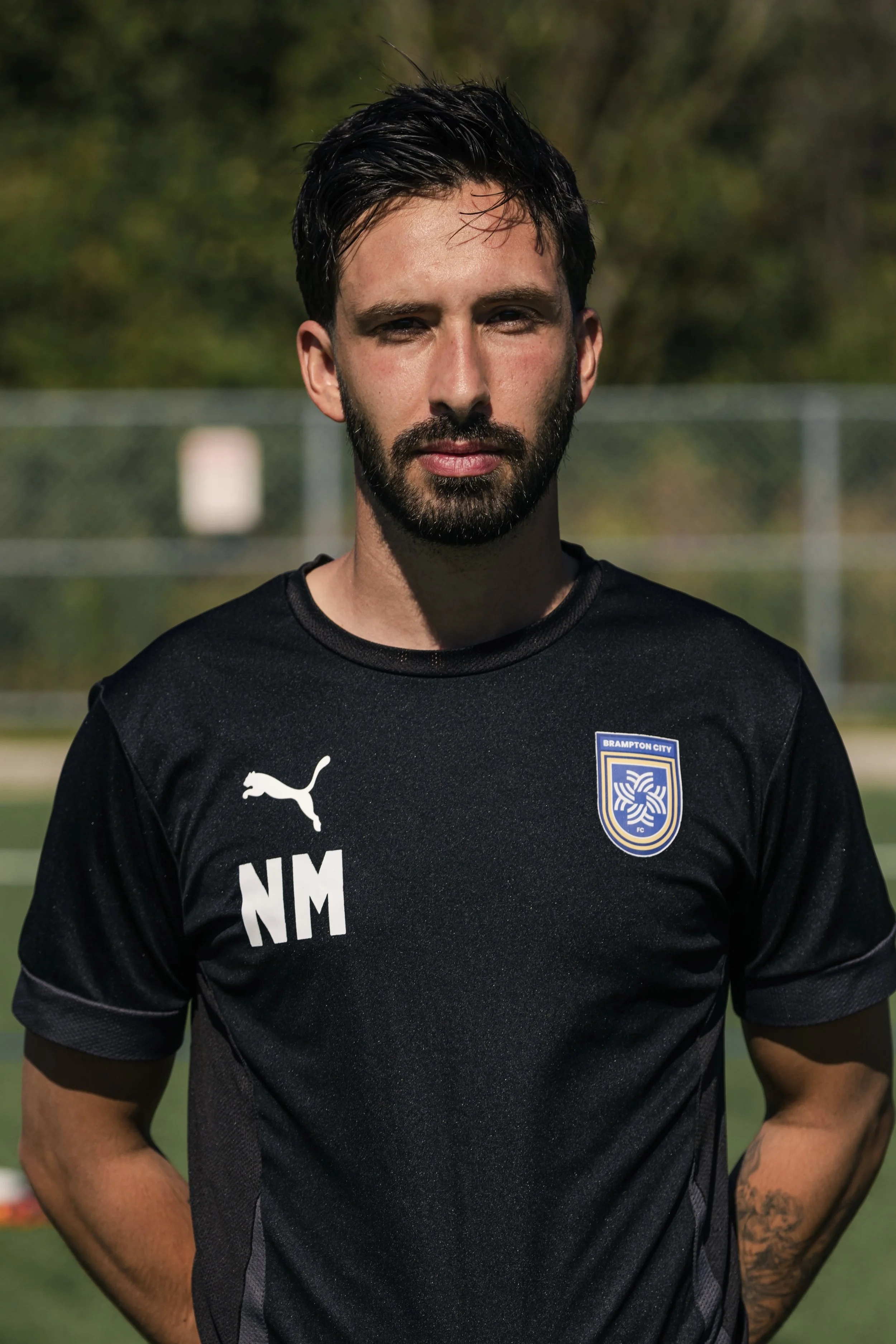 A man with short dark hair and a beard, wearing a black sports jersey with Puma logo and an emblem that reads Brampton City FC, standing outdoors with green trees in the background.