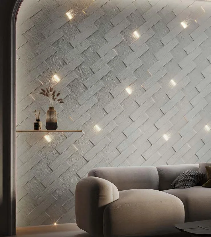 A modern living room with a textured white tile wall, illuminated by small ceiling lights. A light gray plush sofa with a patterned pillow is in front, and a floating shelf with decorative vases and dried flowers is mounted on the wall.