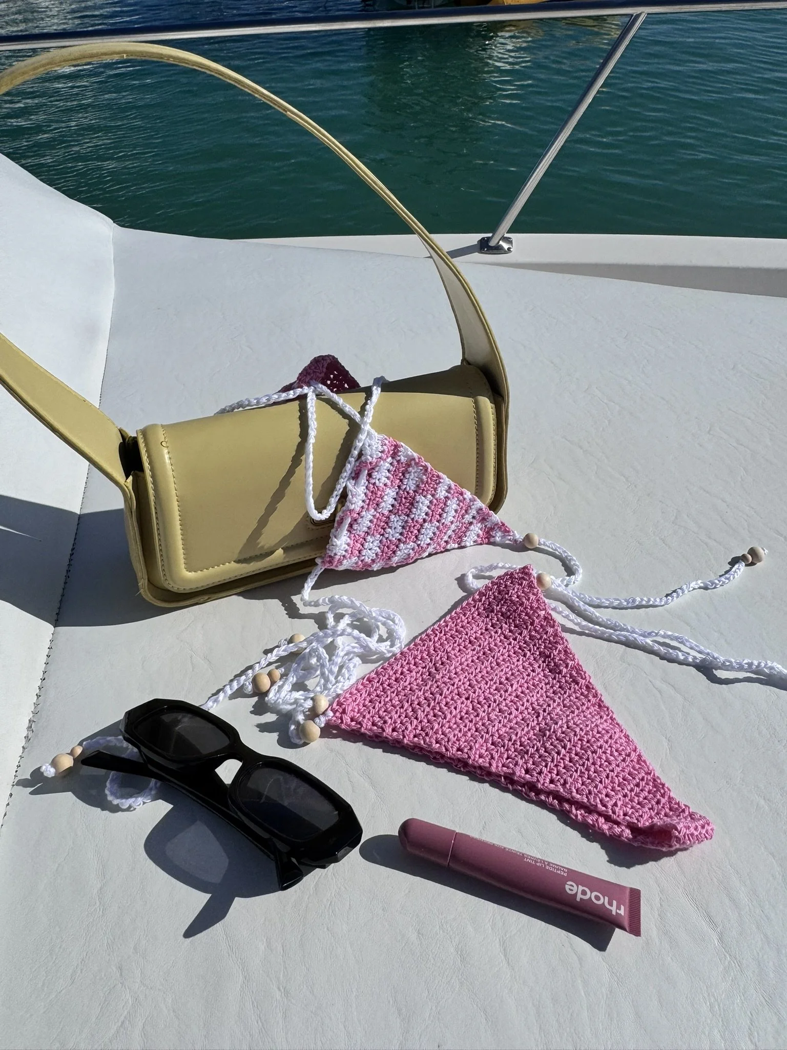 A boat surface with a yellow purse, crocheted pink and white swimsuit, black sunglasses, and a pink lip gloss against the water and sky background.