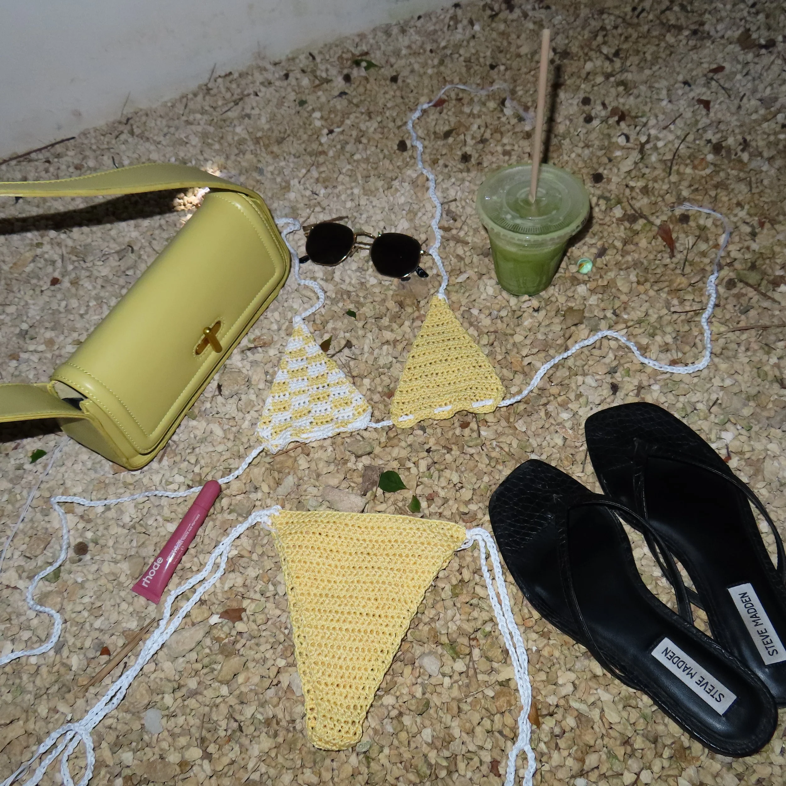 Yellow handbag, black sunglasses, crochet bikini top and bottom, pink lipstick, black Steve Madden sandals, and a green smoothie on gravel ground.
