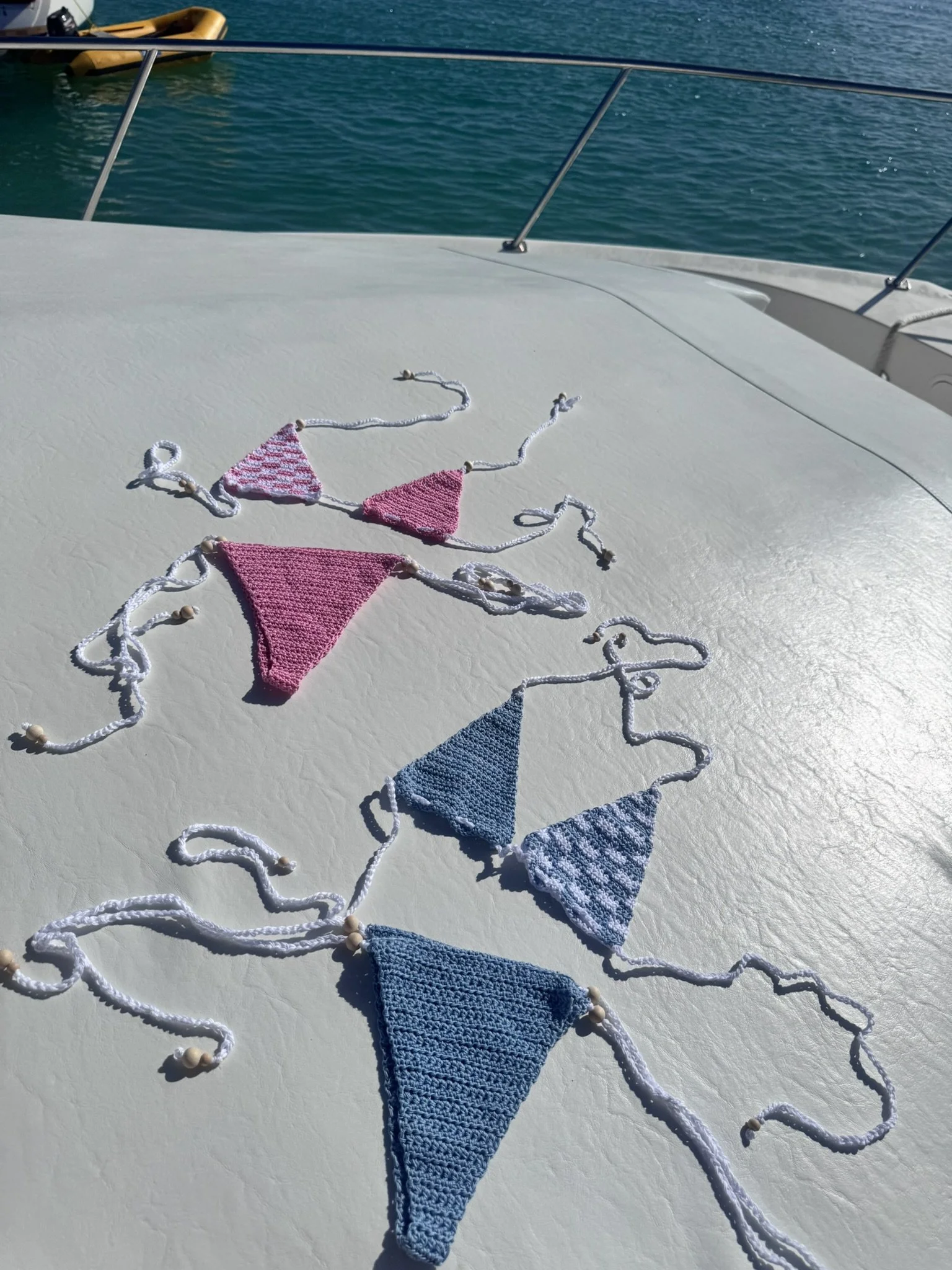Colorful knitted bikini top and string ties laid out on a boat deck, with boats and water in the background.
