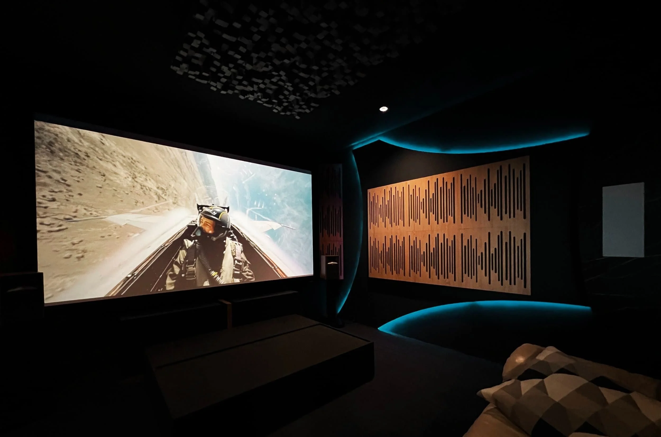 A private home cinema with a large screen showing a jet pilot, acoustic panels, blue LED lighting, and a beige couch.