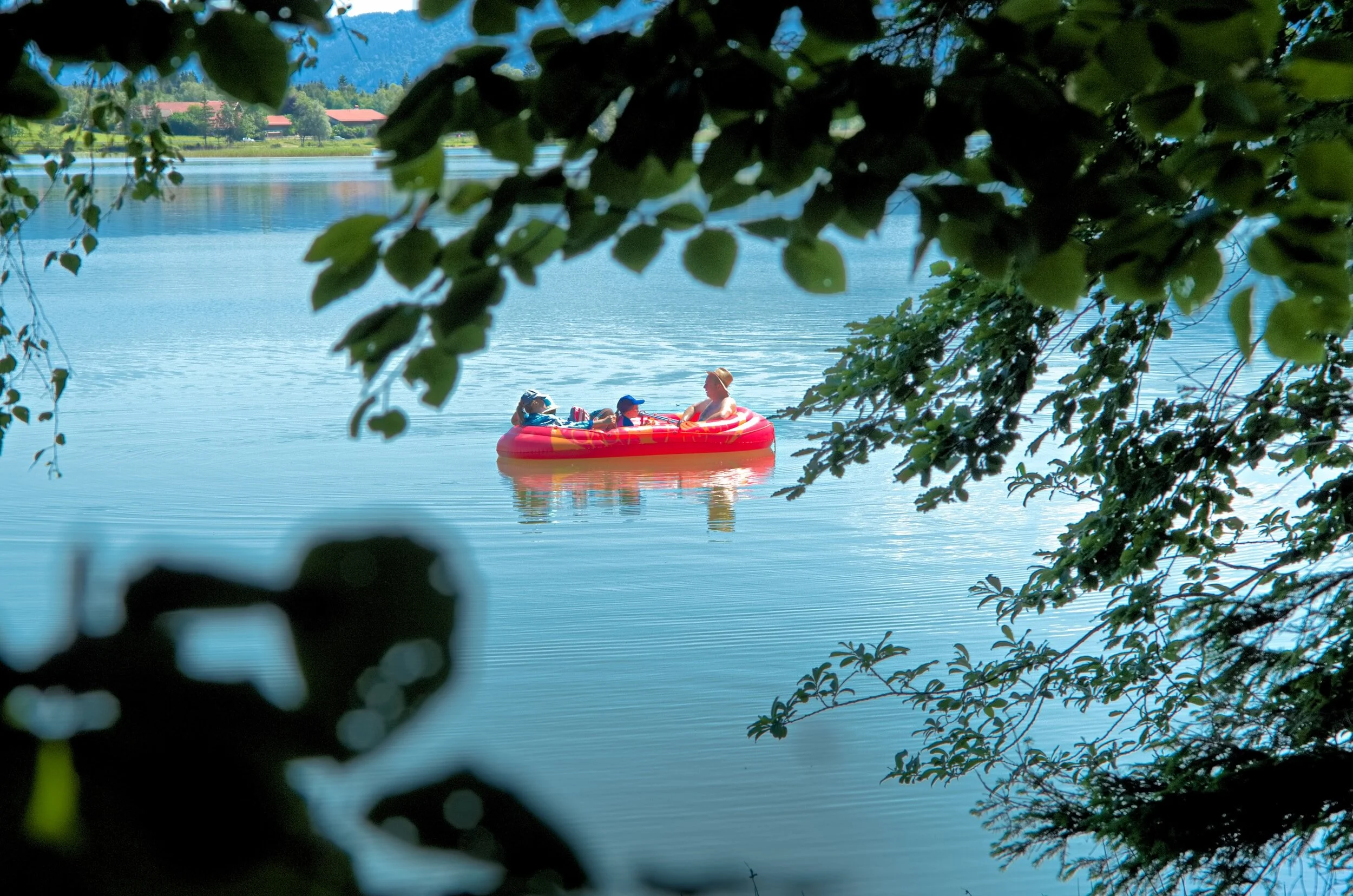 People in a red inflatable boat seen from the shore, floating down the Kettle River towards Christina Lake. One of the many activities to do while camping at Skands.