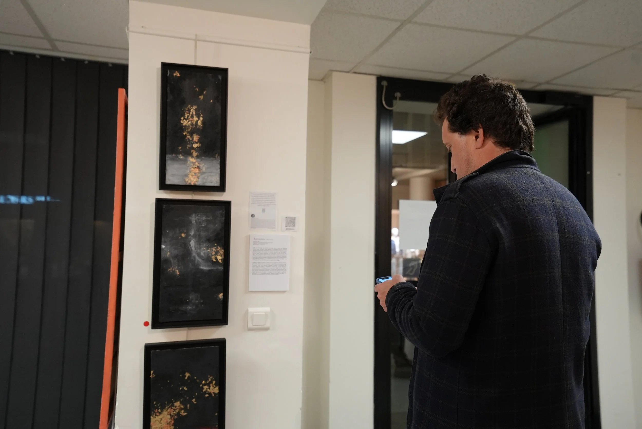 A man in a dark plaid jacket standing in an art gallery, looking at his phone. On the wall in front of him are four vertical framed artworks featuring abstract, dark-colored images with gold accents.
