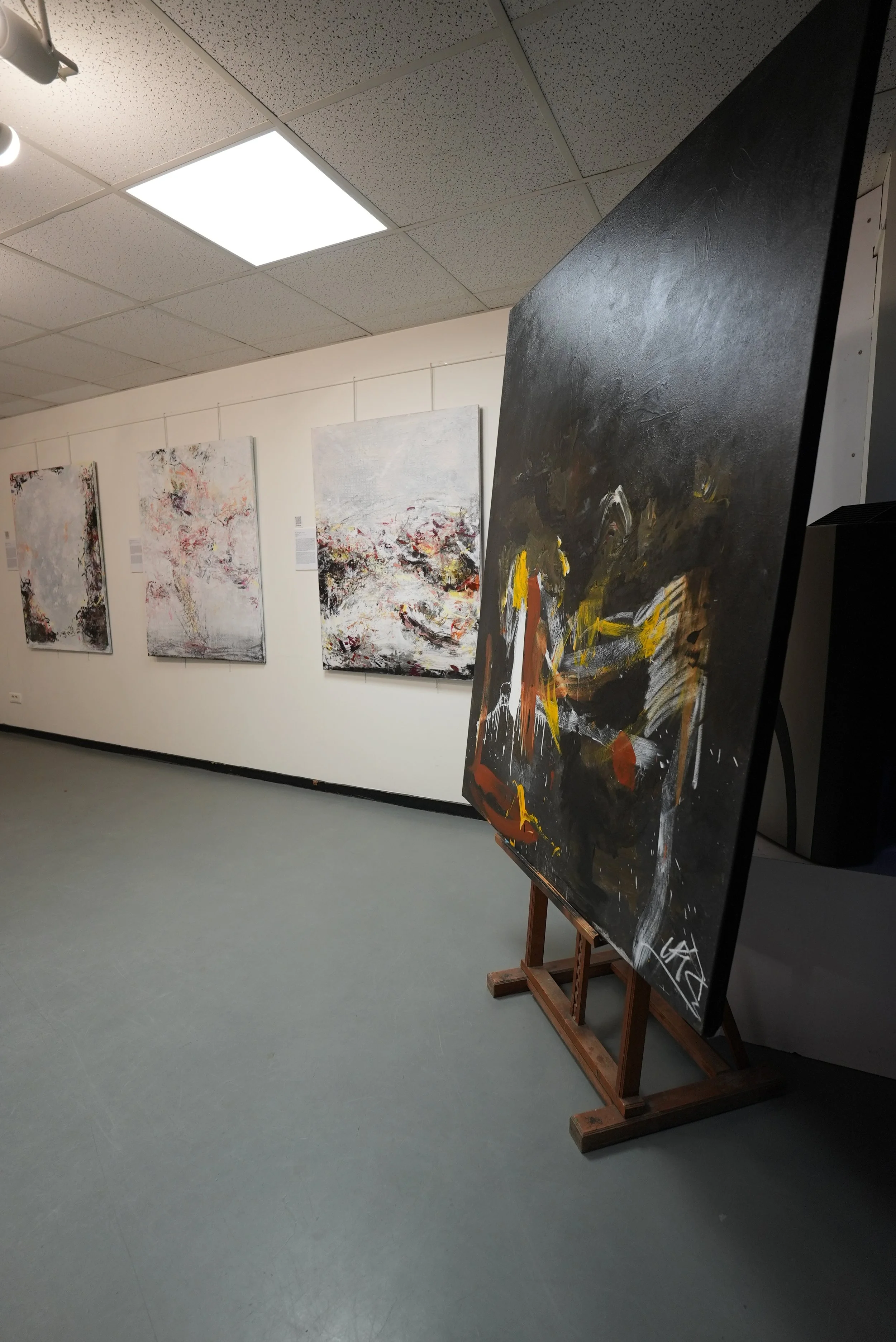 An art gallery with four abstract paintings on the wall, and one painting on an easel in the foreground with a black background and bold brush strokes of yellow, white, and brown.