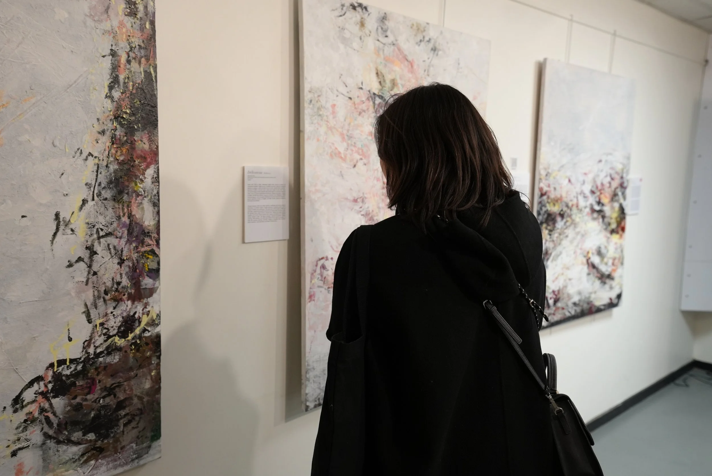 A woman with shoulder-length dark hair observing abstract paintings on a gallery wall, dressed in black, with a black handbag over her shoulder.