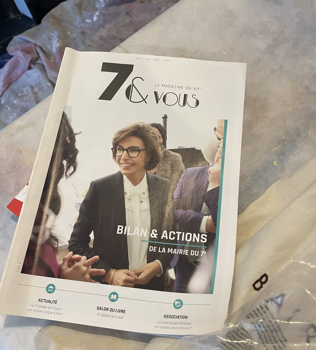 A magazine titled '7 & Vous' with a cover photo of a woman with glasses, smiling, in a business suit, at a meeting or event. The magazine features a headline about the city's 7th arrondissement's action report. It is placed on a table covered with a cloth, with some other items partially visible around it.