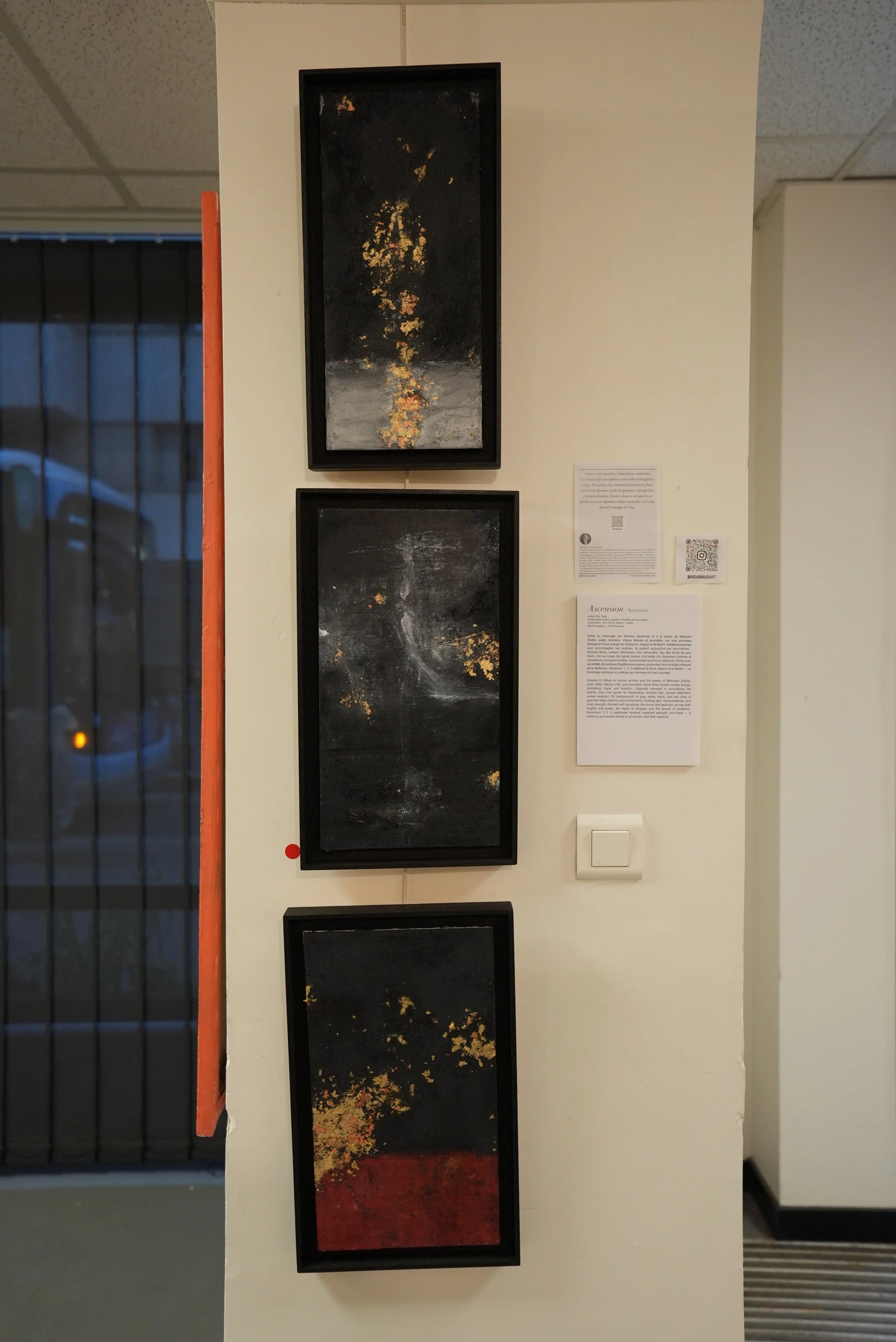 Three abstract paintings with black backgrounds and specks of gold, yellow, and red, hung vertically on a white wall in an art gallery or exhibition space.