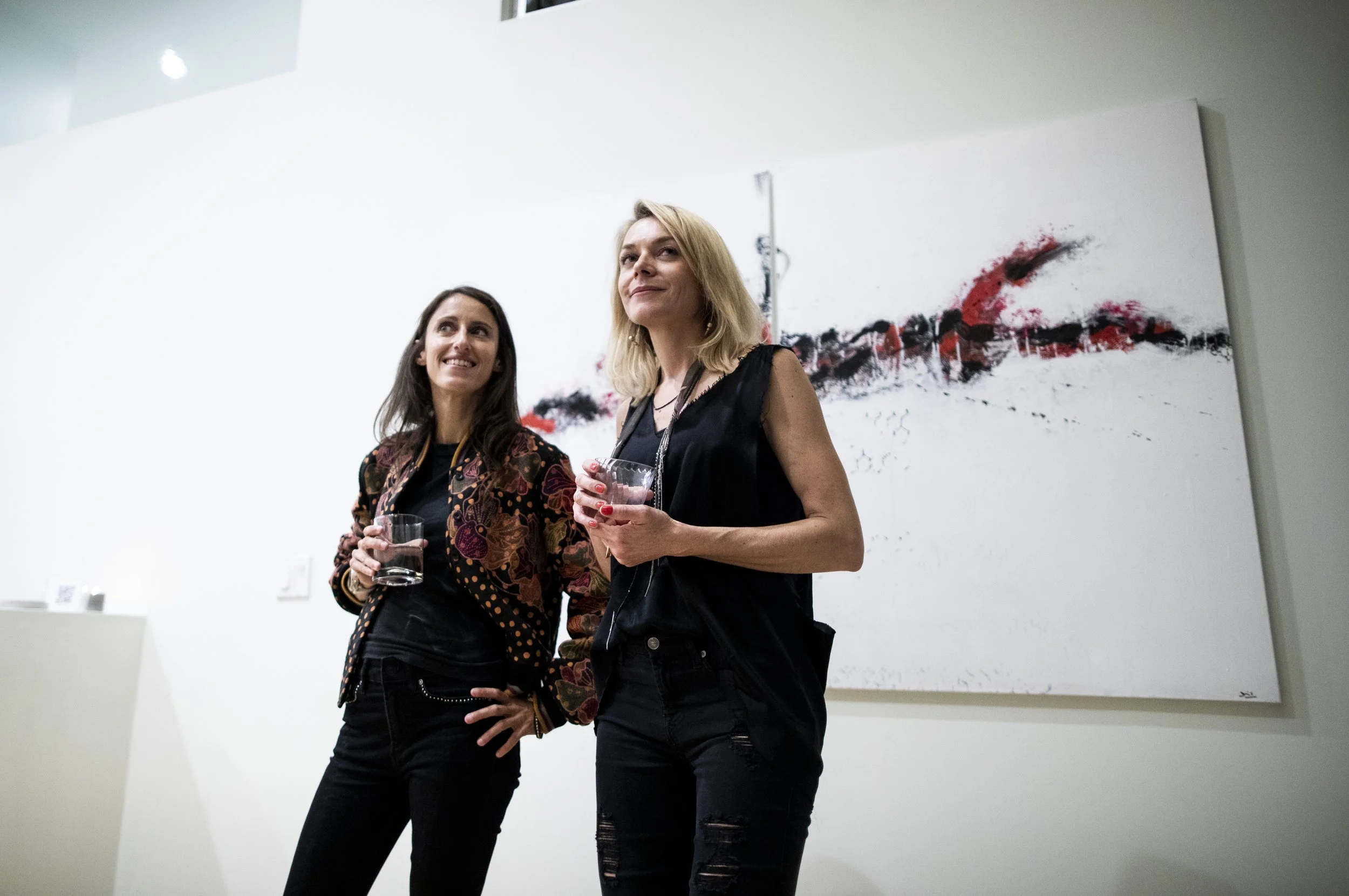 Two women standing in front of abstract artwork at an art gallery, holding drinks, smiling, and looking in different directions. metallica band painting tribute, trujillos