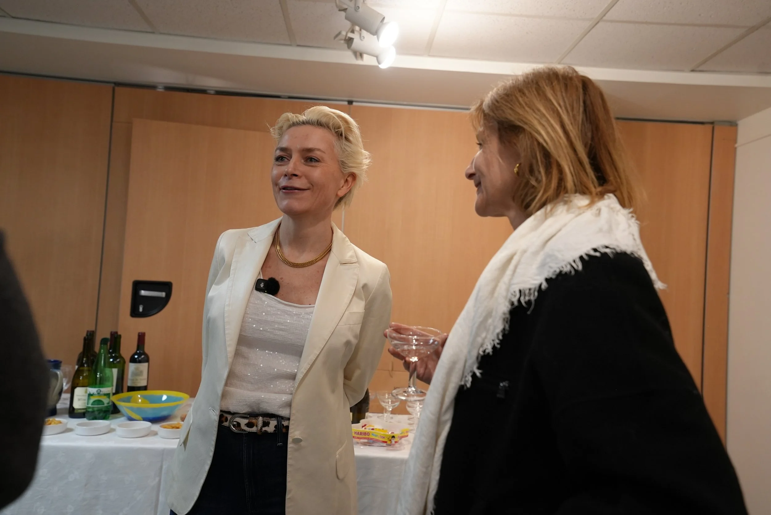Two women engaged in conversation at a social gathering, with a table of snacks and bottles of wine in the background.