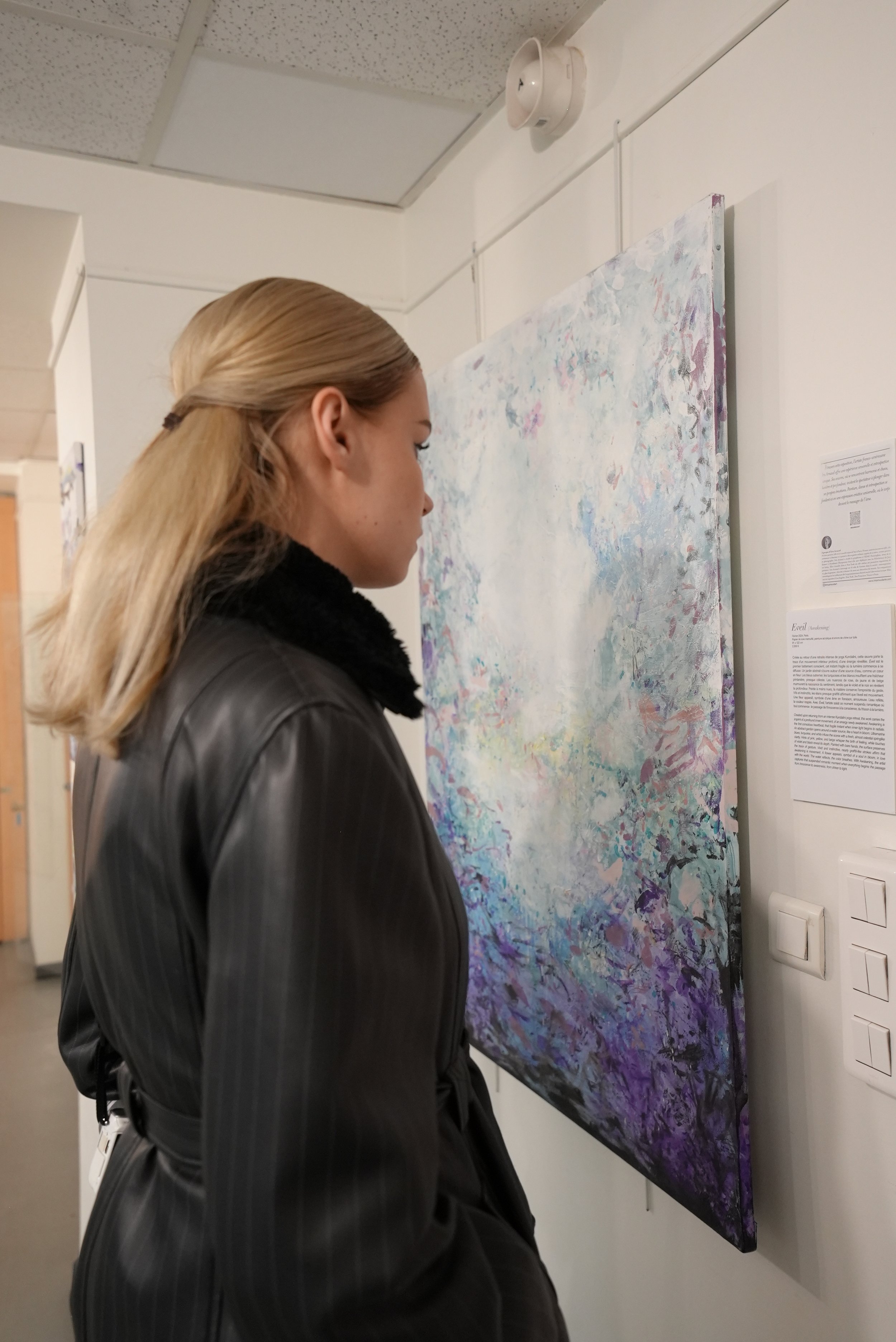 A woman with blonde hair, wearing a black leather coat, looks at an abstract painting with pastel colors on a white gallery wall.