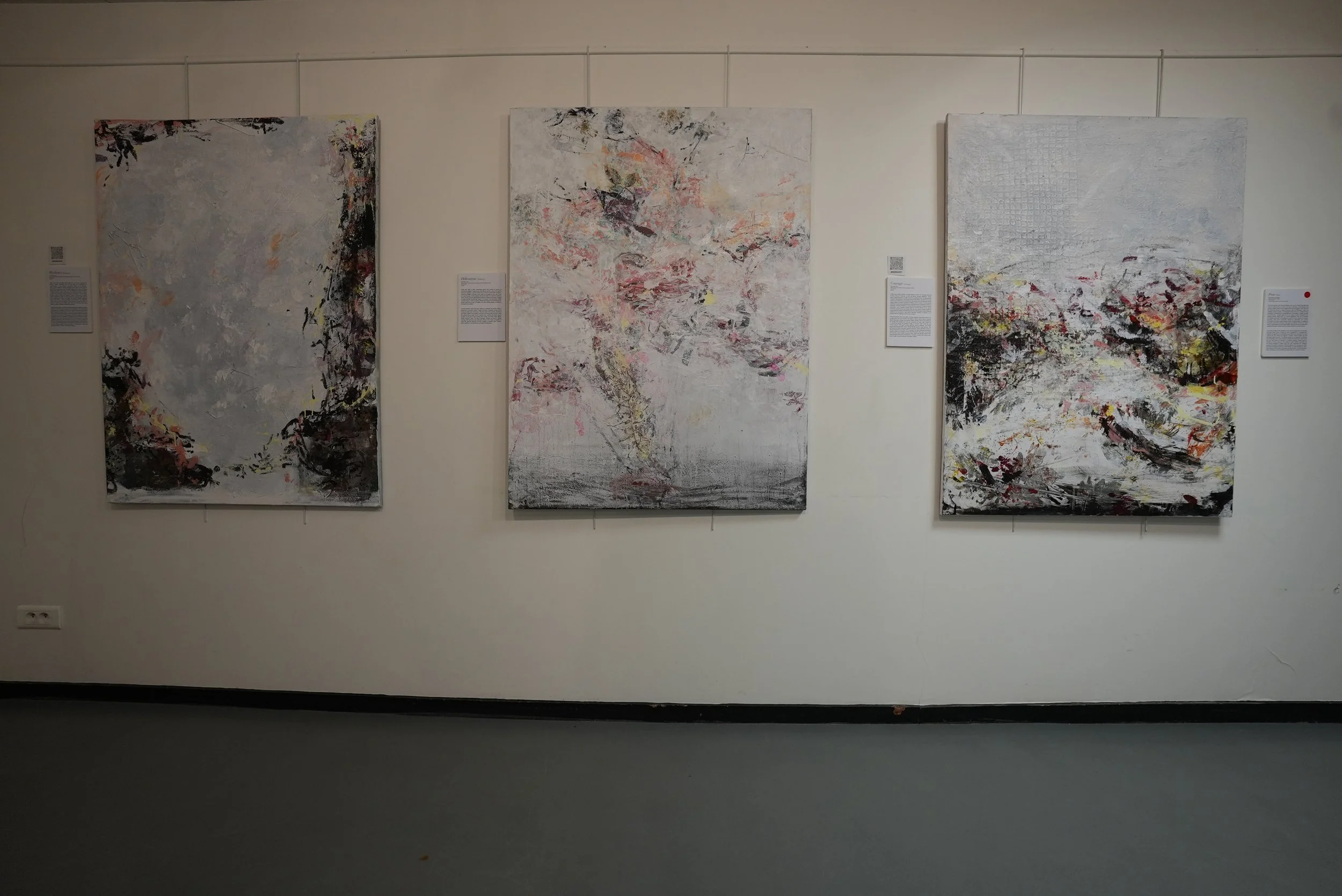 Three abstract paintings hung on a white gallery wall, in Paris France, each with a combination of black, white, pink, and yellow brush strokes.