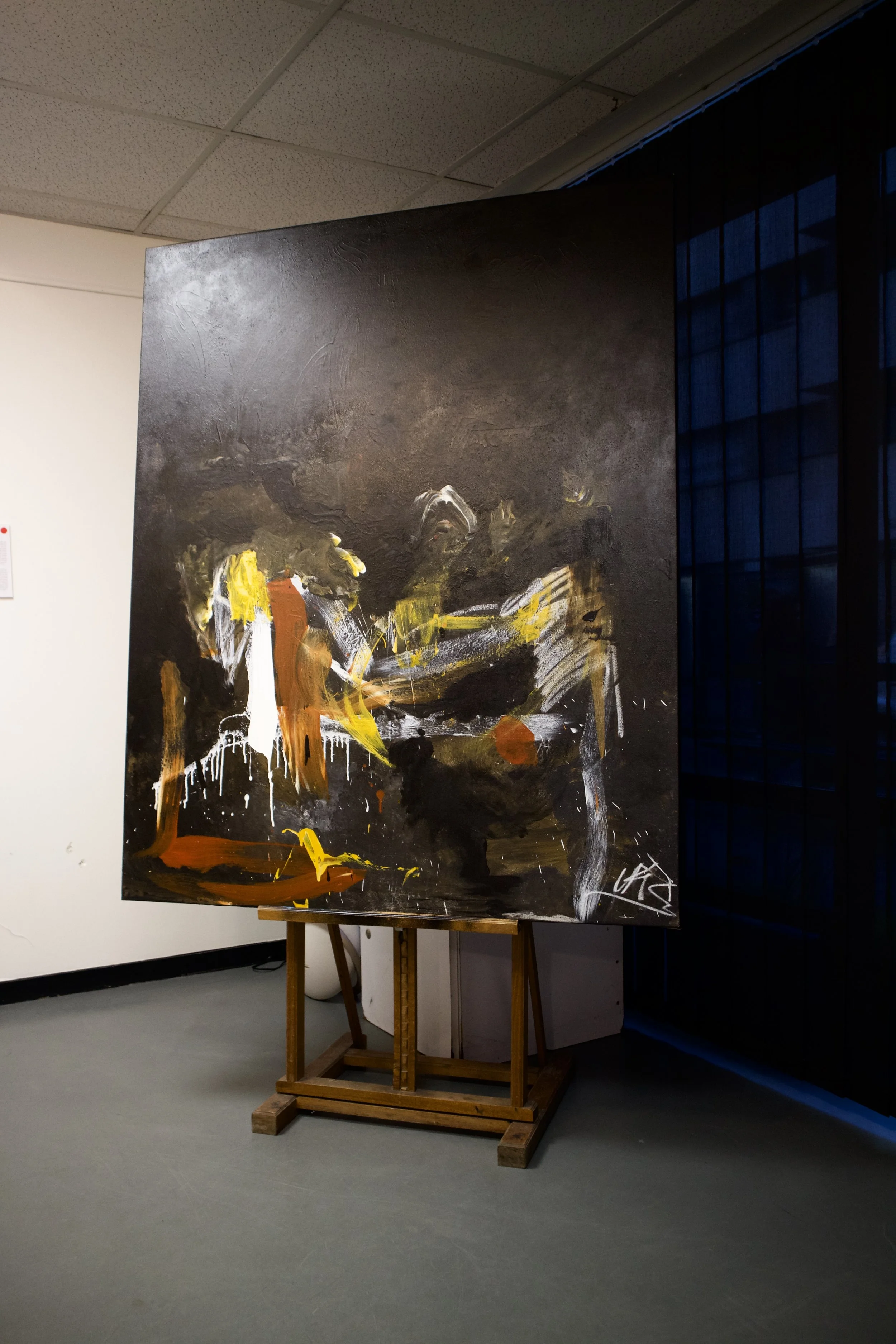 Abstract painting with dark background, yellow, orange, white, and gray brushstrokes displayed on an easel in a gallery.