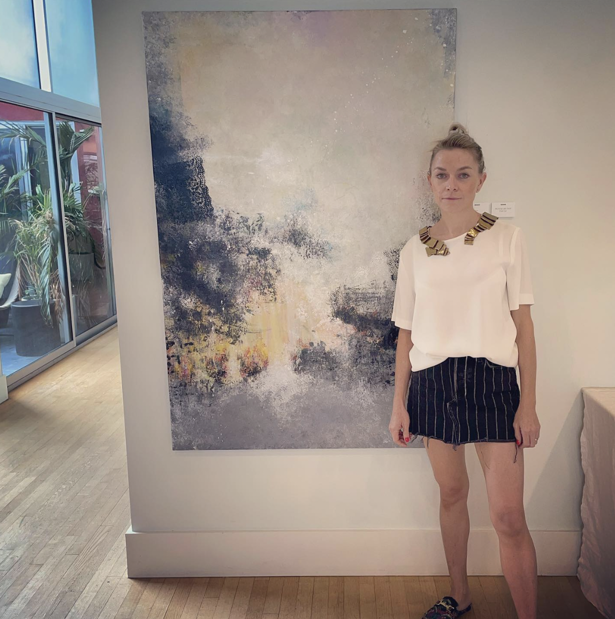 A woman standing next to an abstract painting in a well-lit indoor space. She is wearing a white top with decorative gold embellishments around the neckline, a black striped skirt, and colorful shoes. amy winehouse painting tribute.