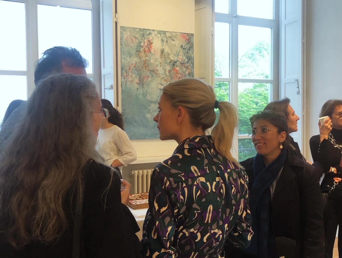 Group of women engaged in conversation at an indoor event, with large windows revealing green trees outside.