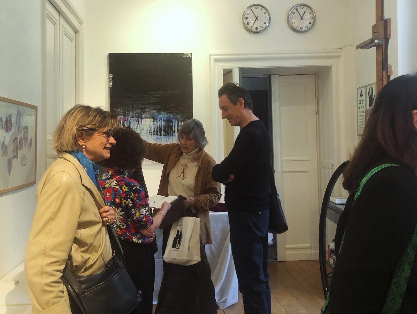 Group of five people talking and interacting in an indoor art gallery or exhibition space, with one person showing a book or pamphlet, and artwork hanging on the walls.