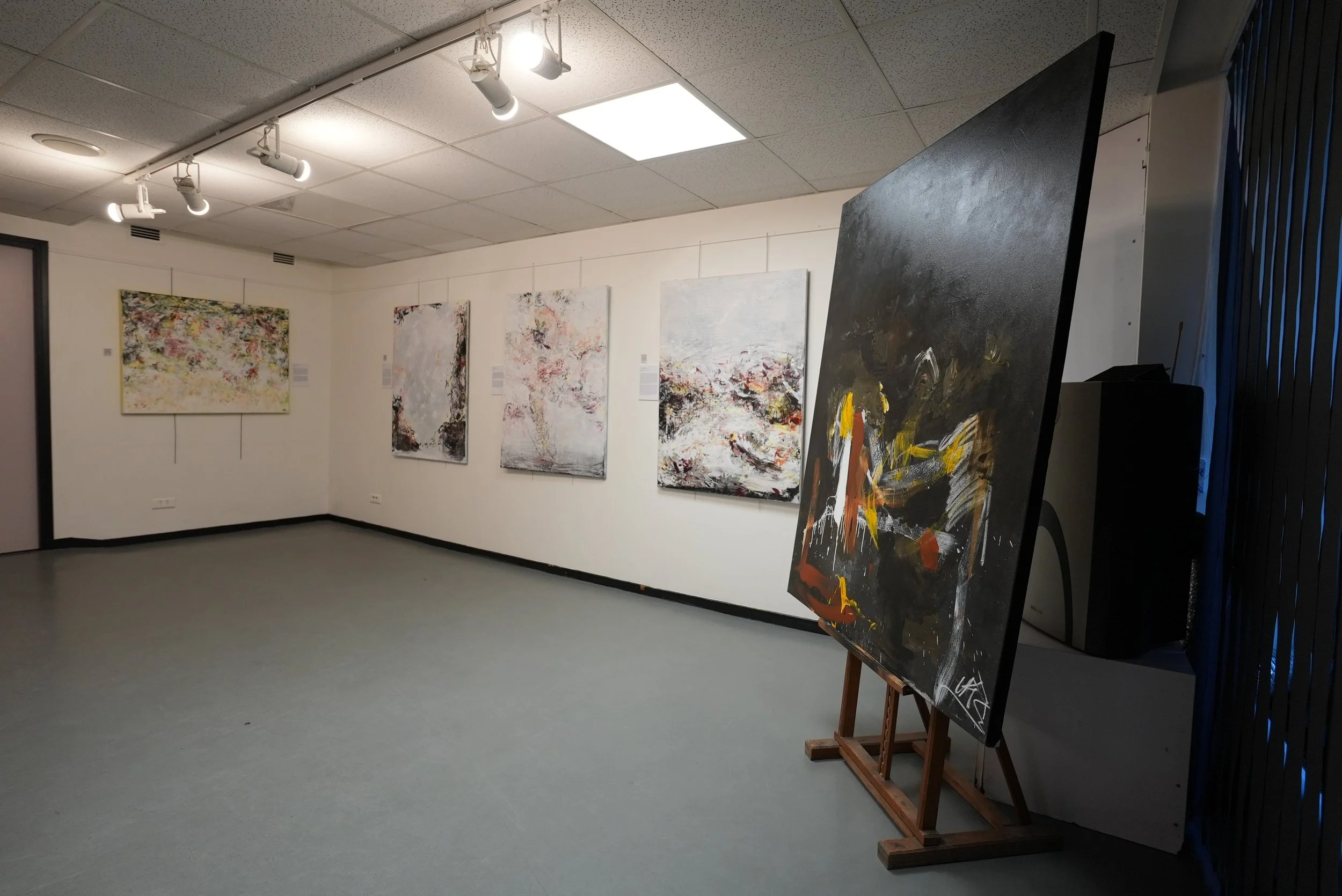 Art gallery with abstract paintings on white walls and a large black abstract painting on an easel. Overhead lighting illuminates the space with a gray floor.