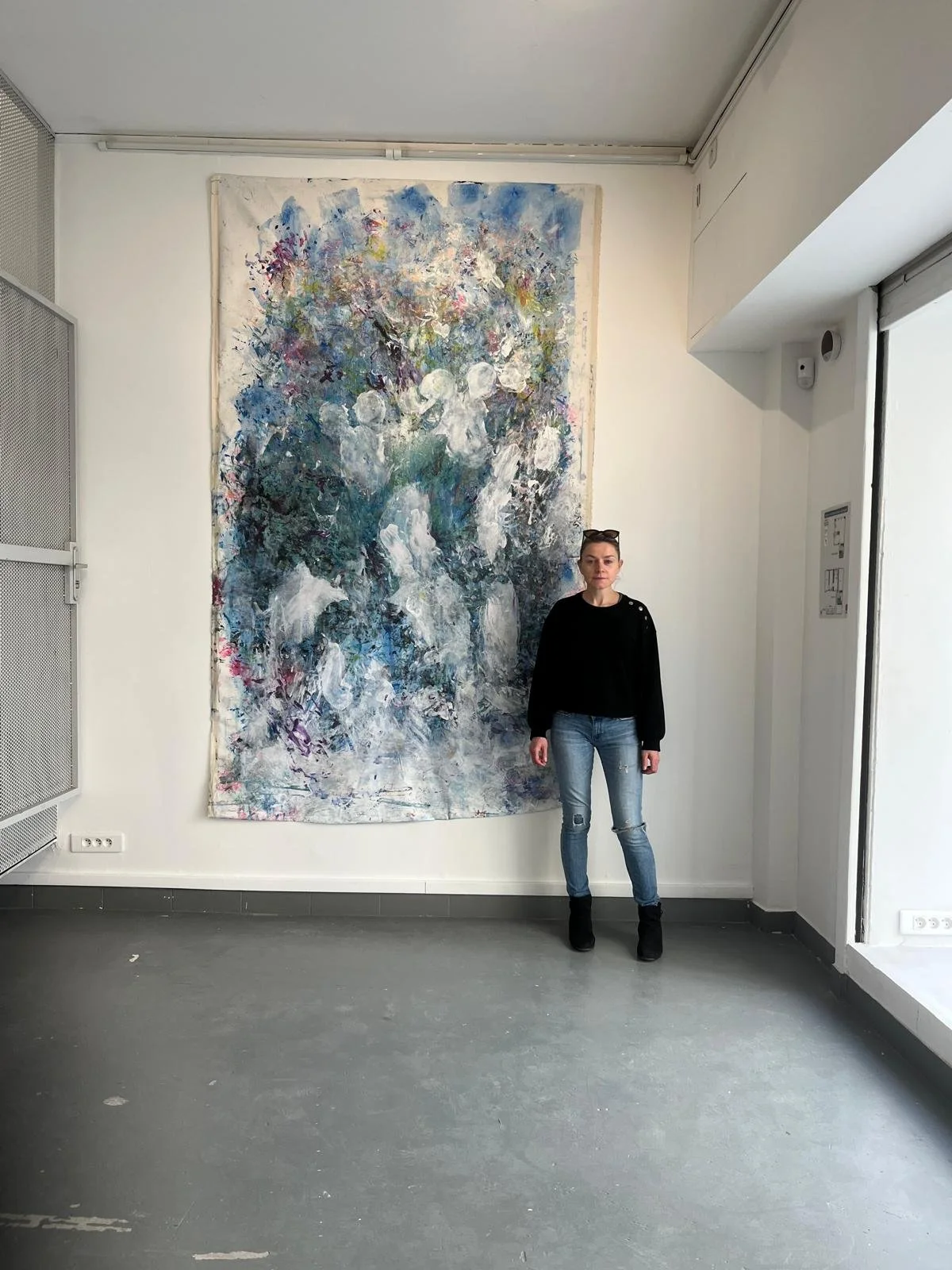 A woman standing in front of a large abstract painting with blue, white, and purple colors inside an art gallery.