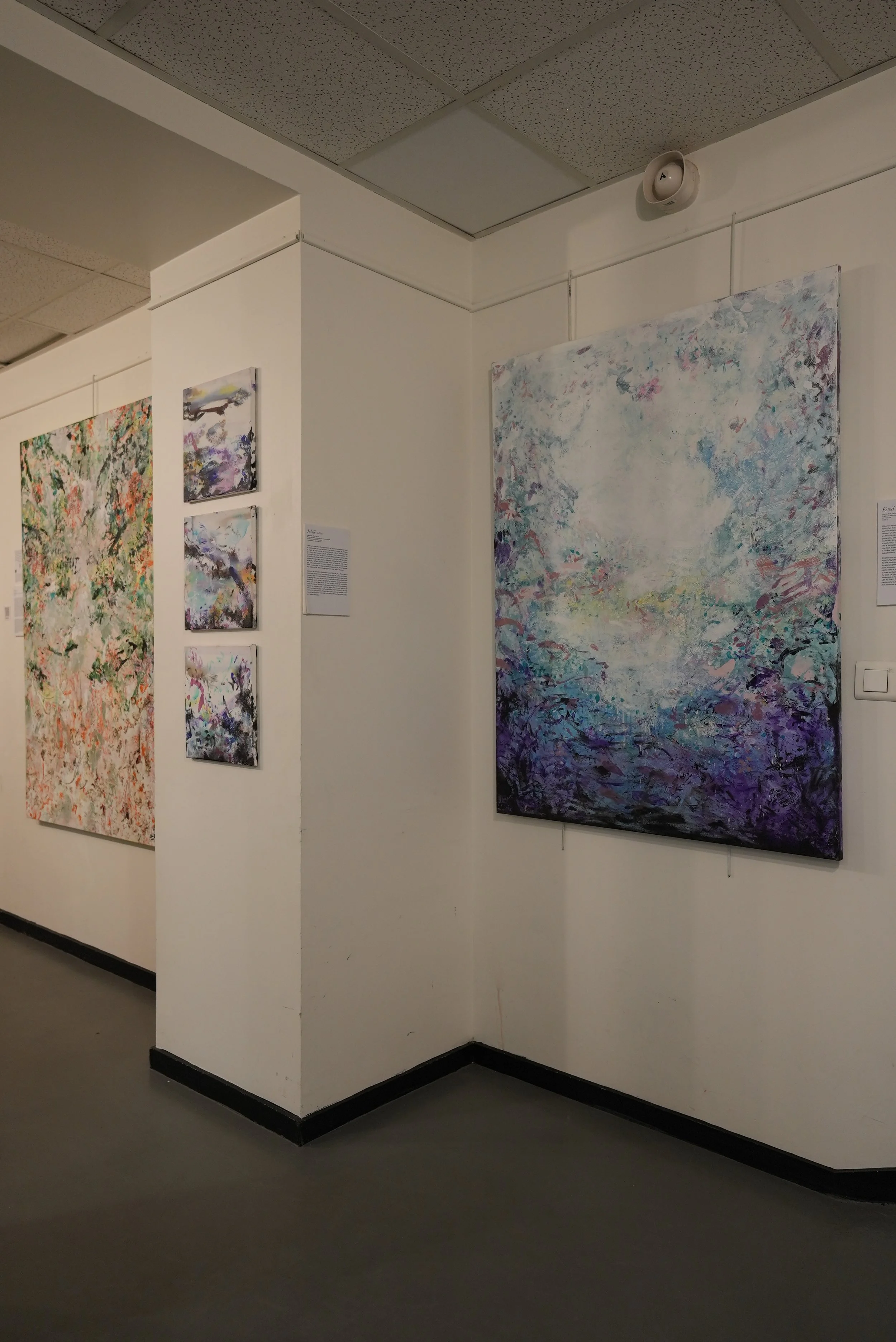 Art gallery with abstract paintings on white walls, including a large piece with shades of blue and purple on the right, and smaller paintings to the left.