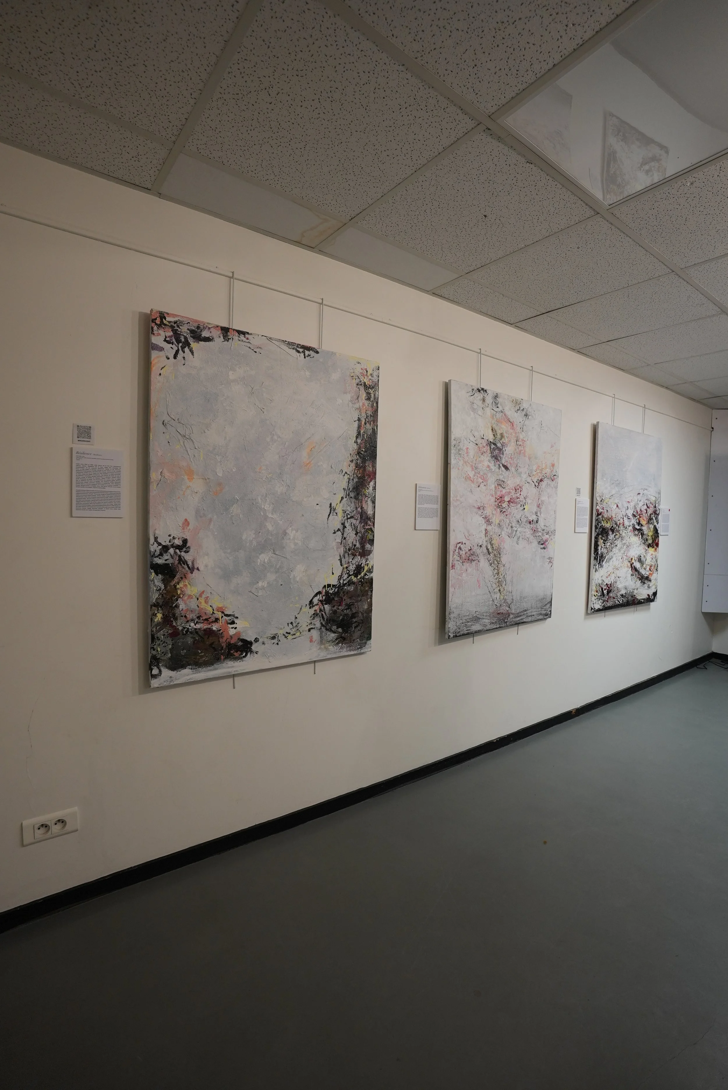 An art gallery wall in Paris, France, displaying three abstract paintings with light colors and textured surfaces, accompanied by informational placards.