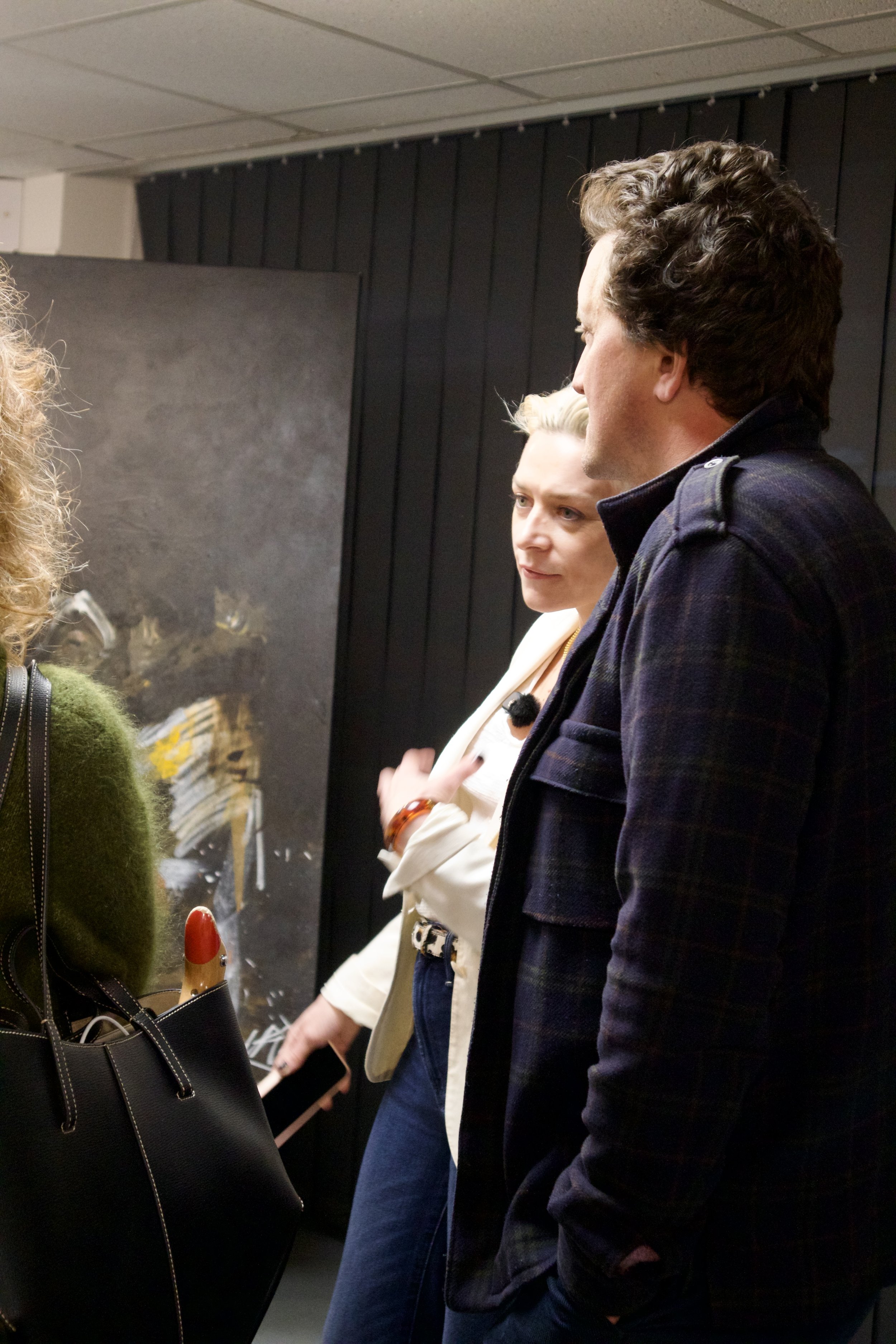 Three people having a conversation indoors, with dark vertical paneling behind them and a blurry painting on the wall.