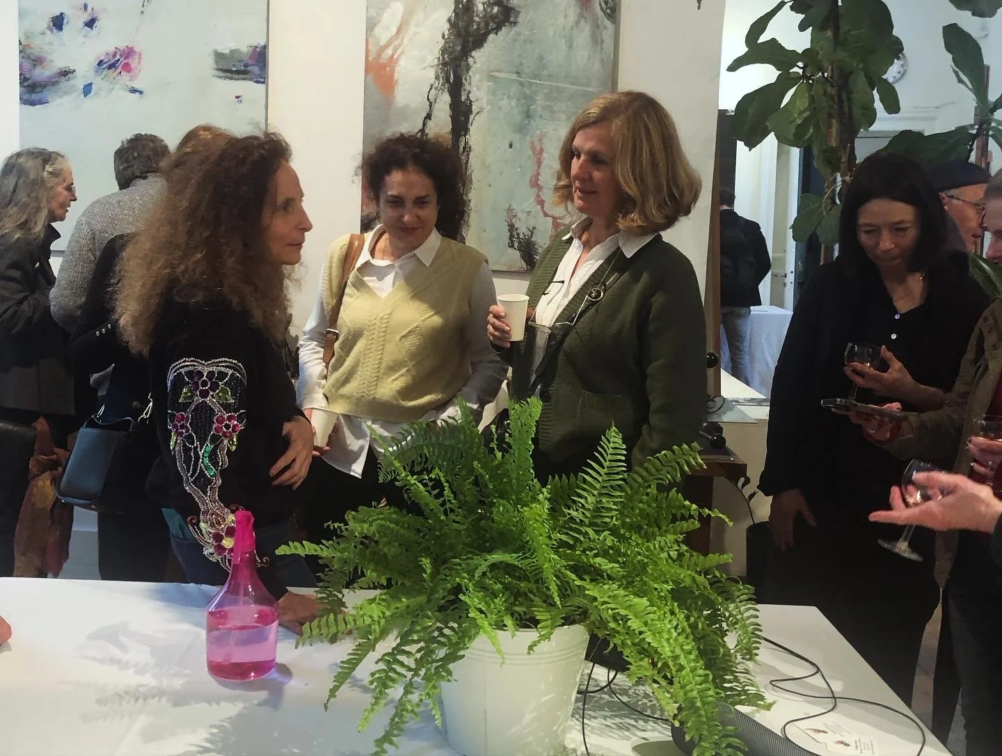 Women engaged in conversation at a social gathering, with a potted fern and a pink spray bottle on a white table in the foreground.