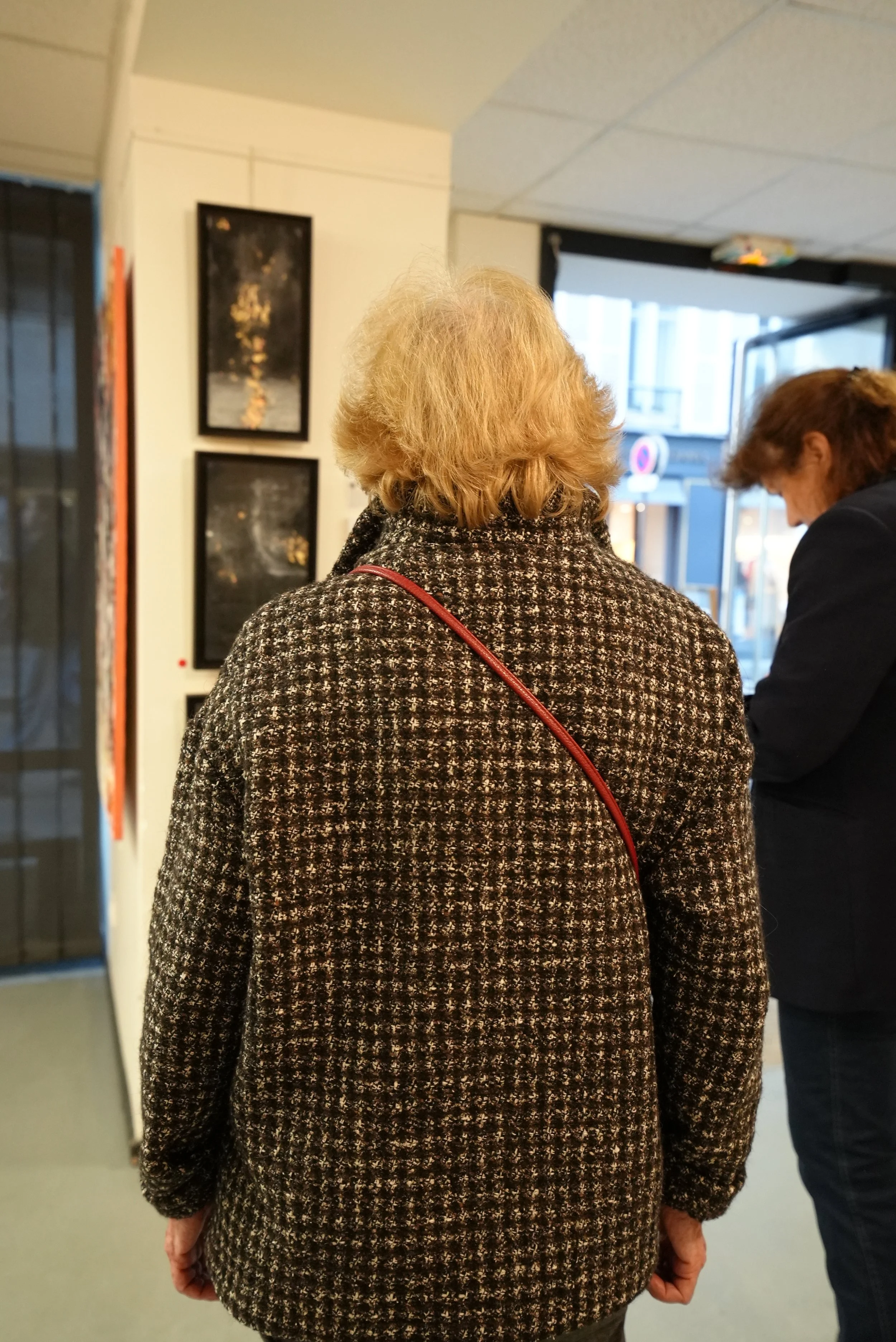Back view of an elderly woman with blonde hair wearing a patterned coat and a red crossbody bag, standing indoors.