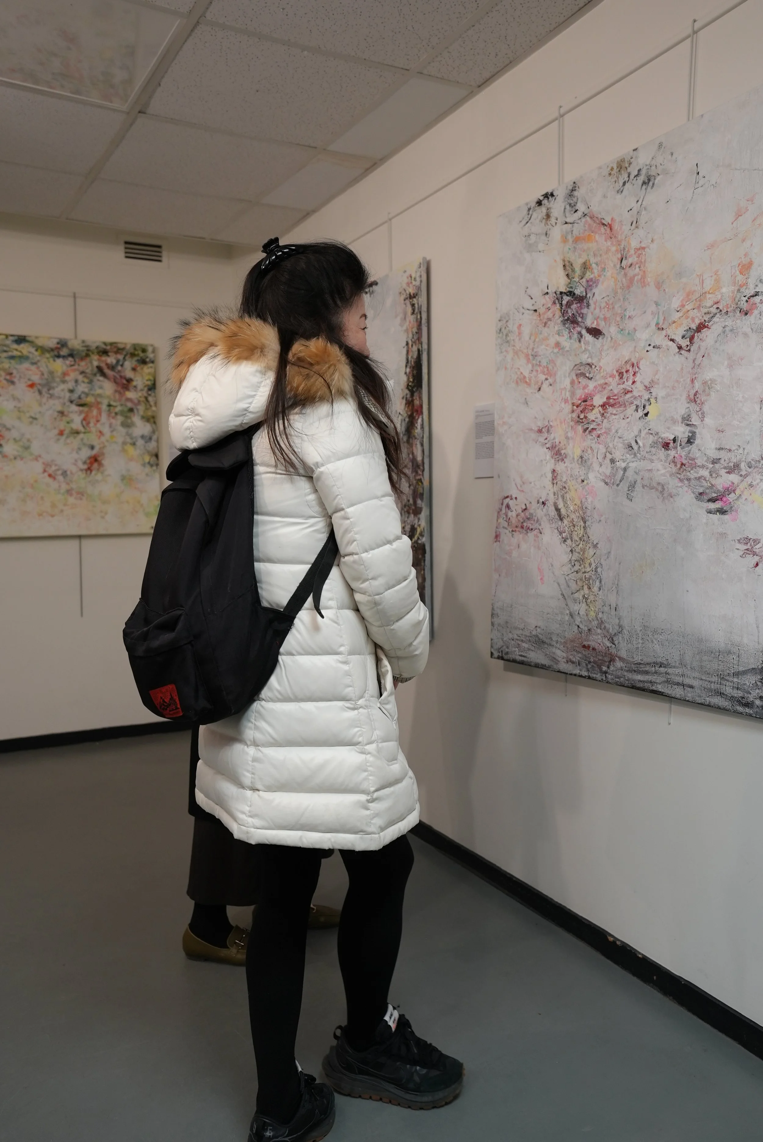 A woman with dark hair wearing a white puffy jacket with a fur-lined hood and a black backpack, observing abstract paintings in an art gallery.