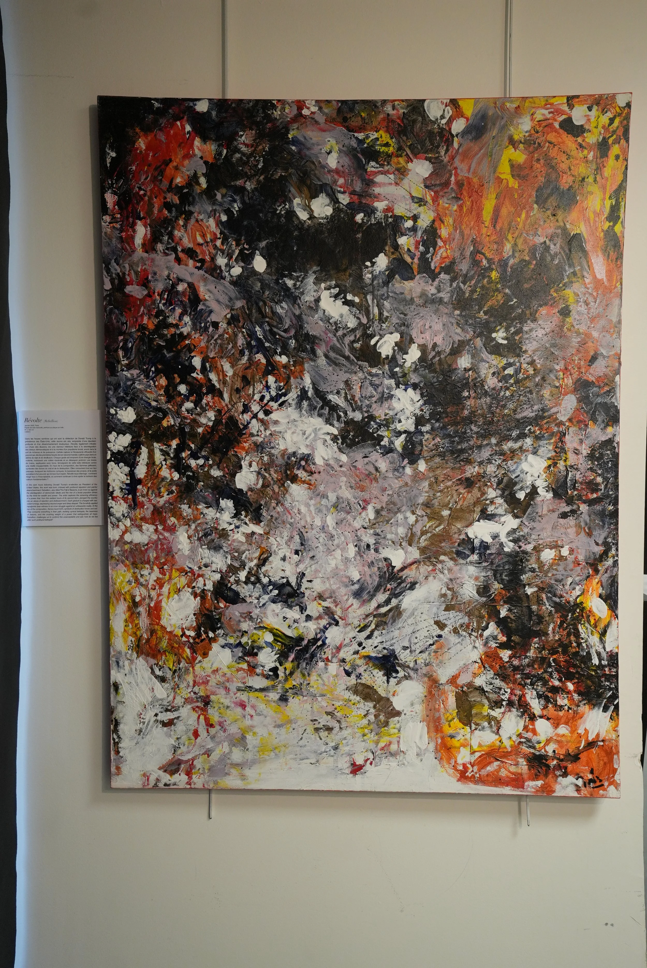 Abstract expressionist painting with black, white, red, orange, yellow, and pink brushstrokes displayed on a white wall, with a descriptive plaque to the left.