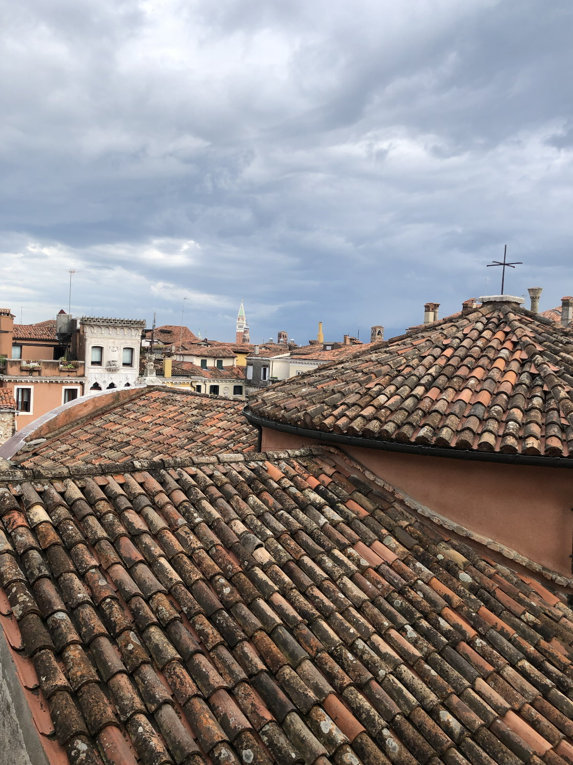 Rooftops of buildings with weathered clay tiles, some moss and lichen, in a European cityscape under cloudy sky with dark clouds and a tower with a green spire in the distance.
