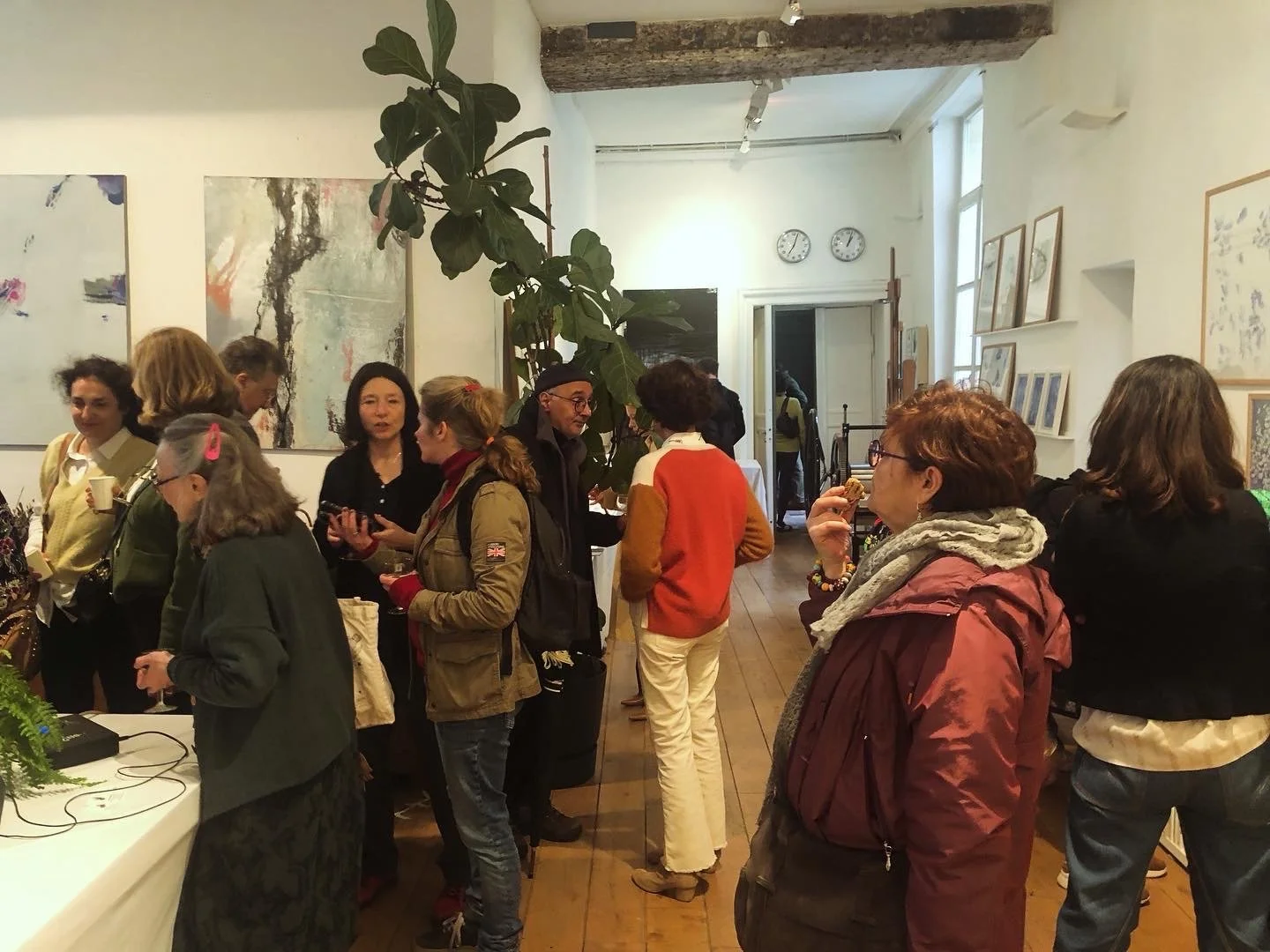People gathered inside an art gallery, mingling and talking, with artwork hanging on the walls and a large plant in the center of the room.