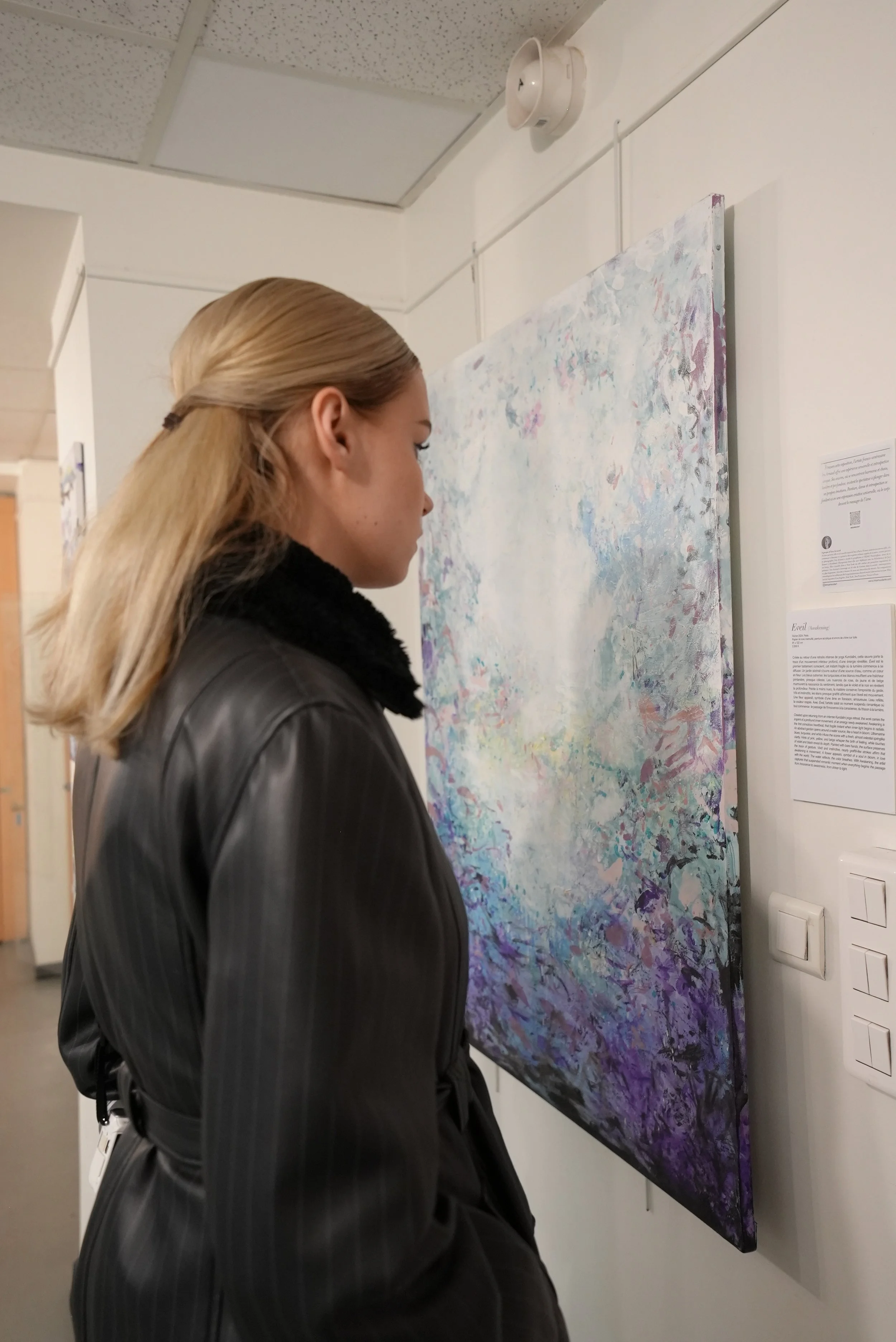 A woman with blonde hair wearing a black pinstripe coat looking at an abstract painting on a gallery wall.