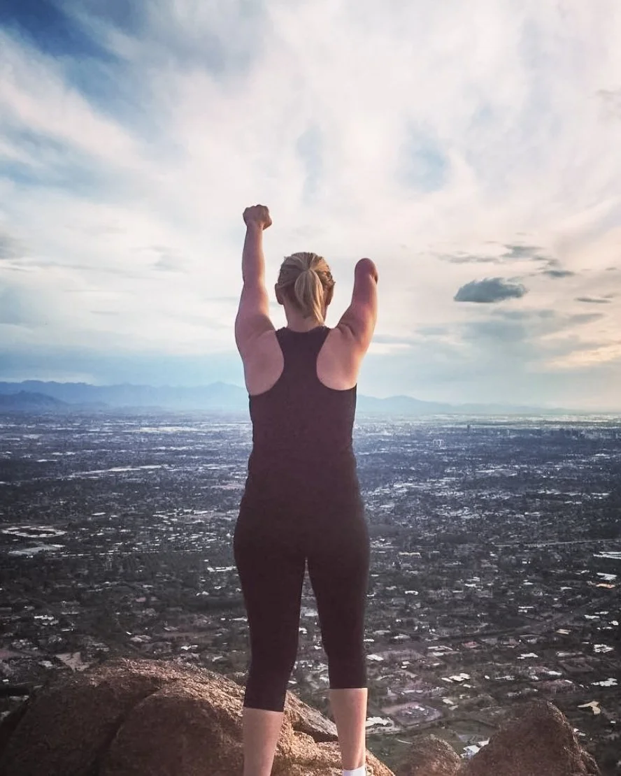 Carrie Christiansen, LPC, Arizona telehealth therapist specializing in anxiety, ADHD, and insomnia, standing on a mountaintop with arms raised overlooking a cityscape.