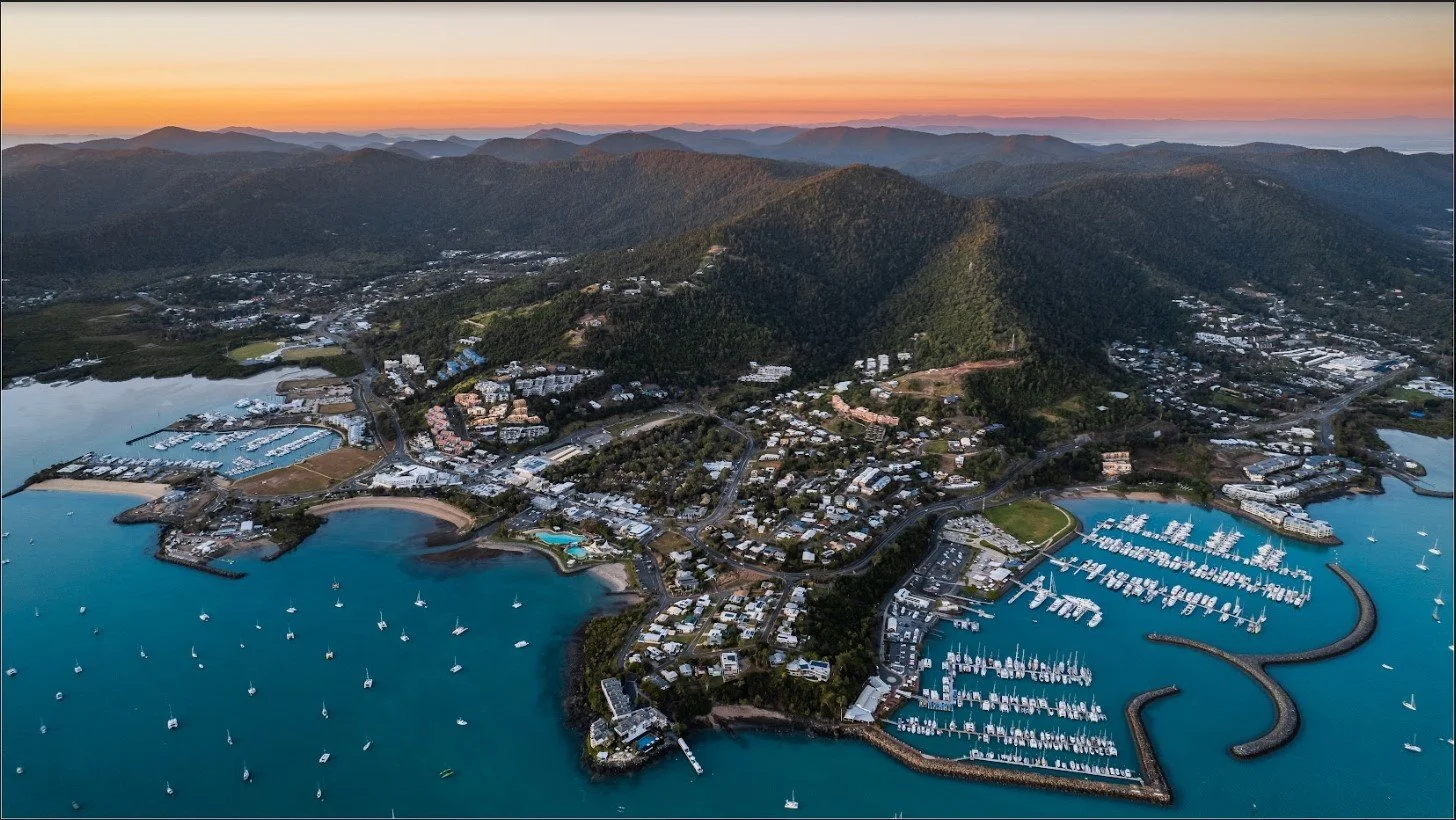 Aerial view of a coastal town with marina and harbor filled with sailboats, surrounded by green mountains at sunset.