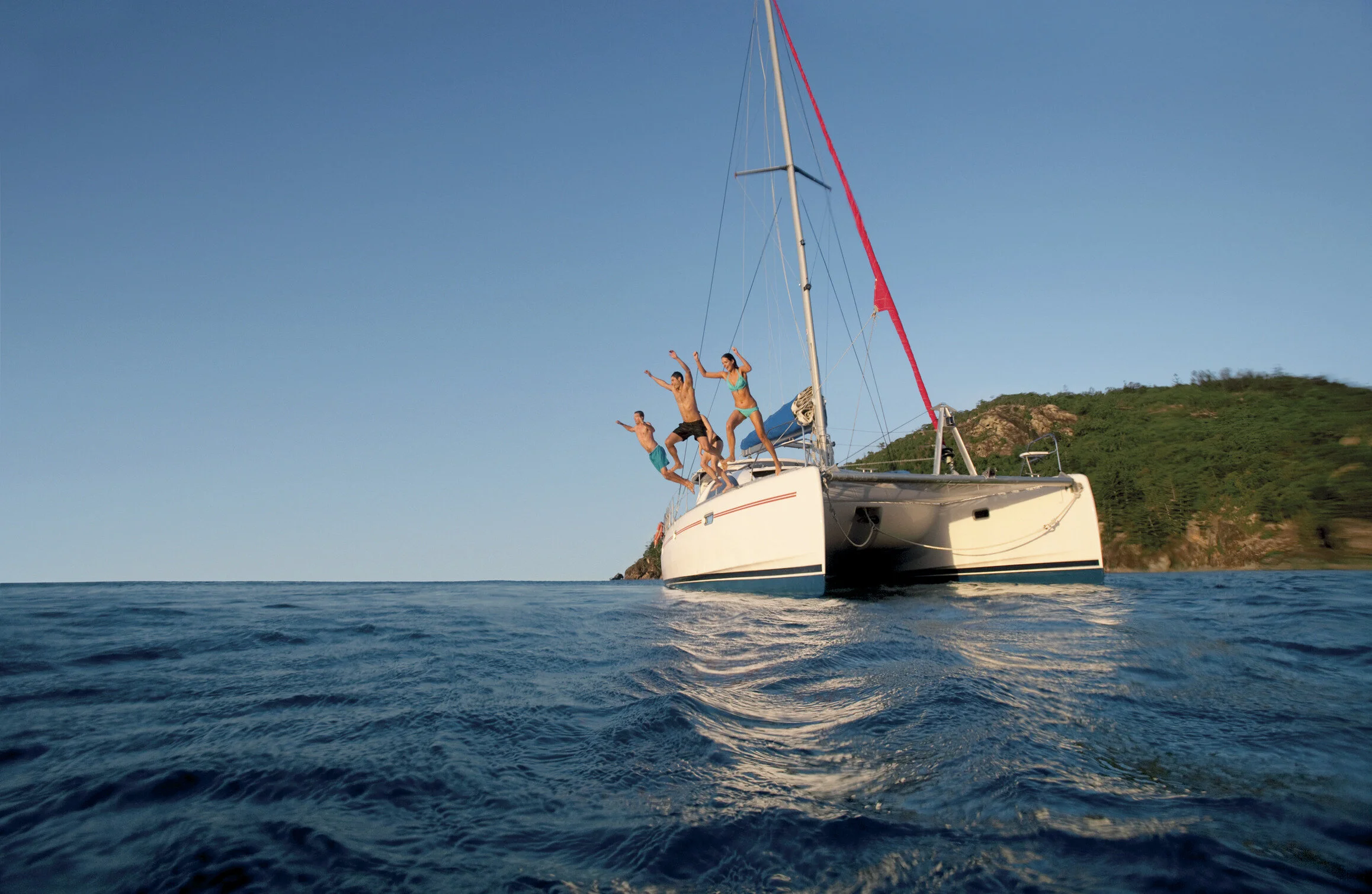 A sailboat with four people on board, standing on the bow, celebrating or enjoying life, with a coastal green hillside in the background and calm blue waters.