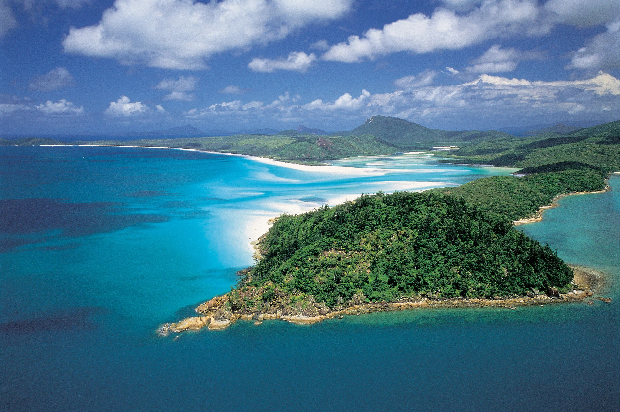 Aerial view of airlie beach coastline with turquoise ocean waters, white sandy beaches, lush green hills, and a partly cloudy sky. Whitsunday Skyway