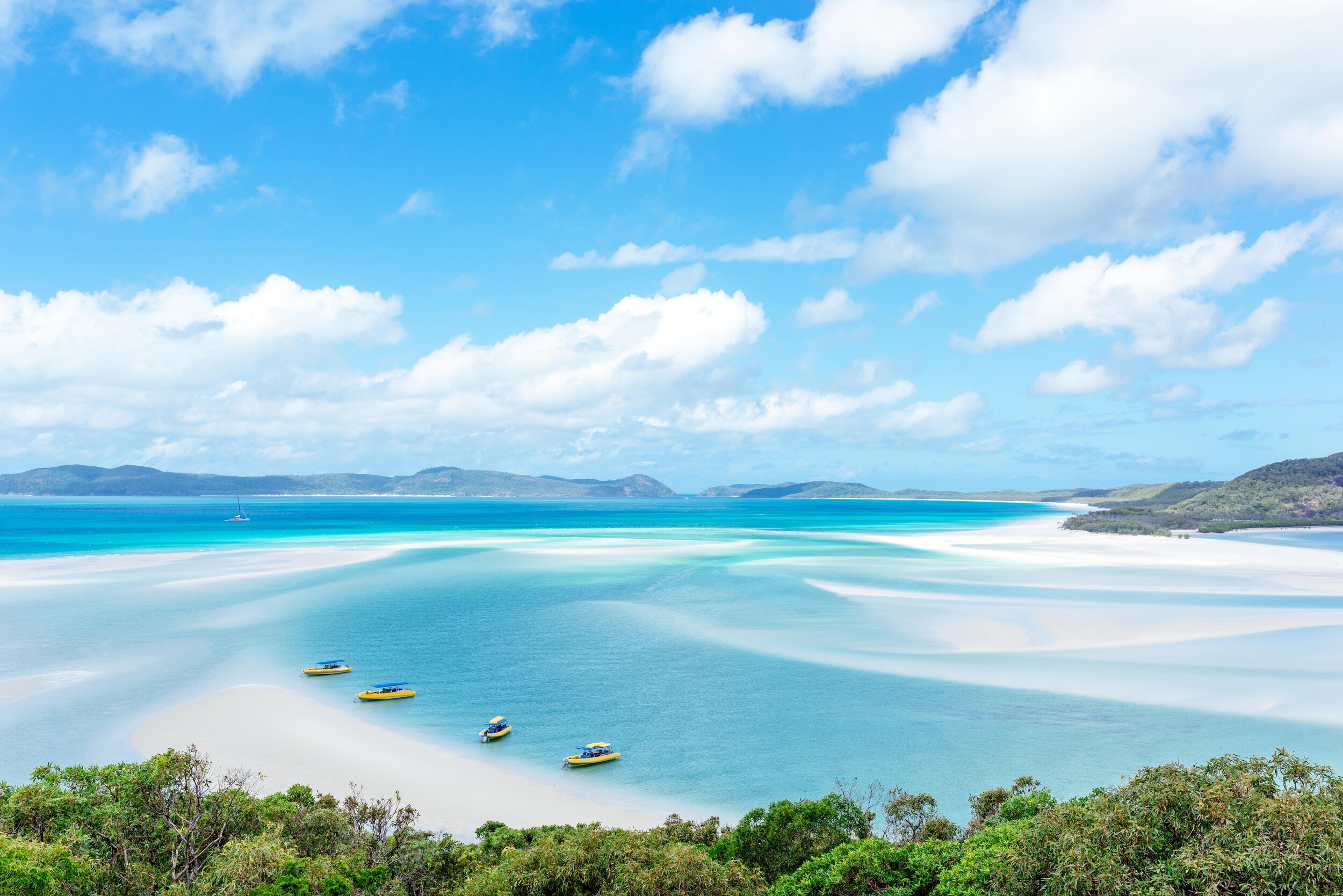 A scenic beach with turquoise waters, white sand, and boats anchored near the shore. Green foliage in the foreground and distant landforms in the background under a partly cloudy sky.