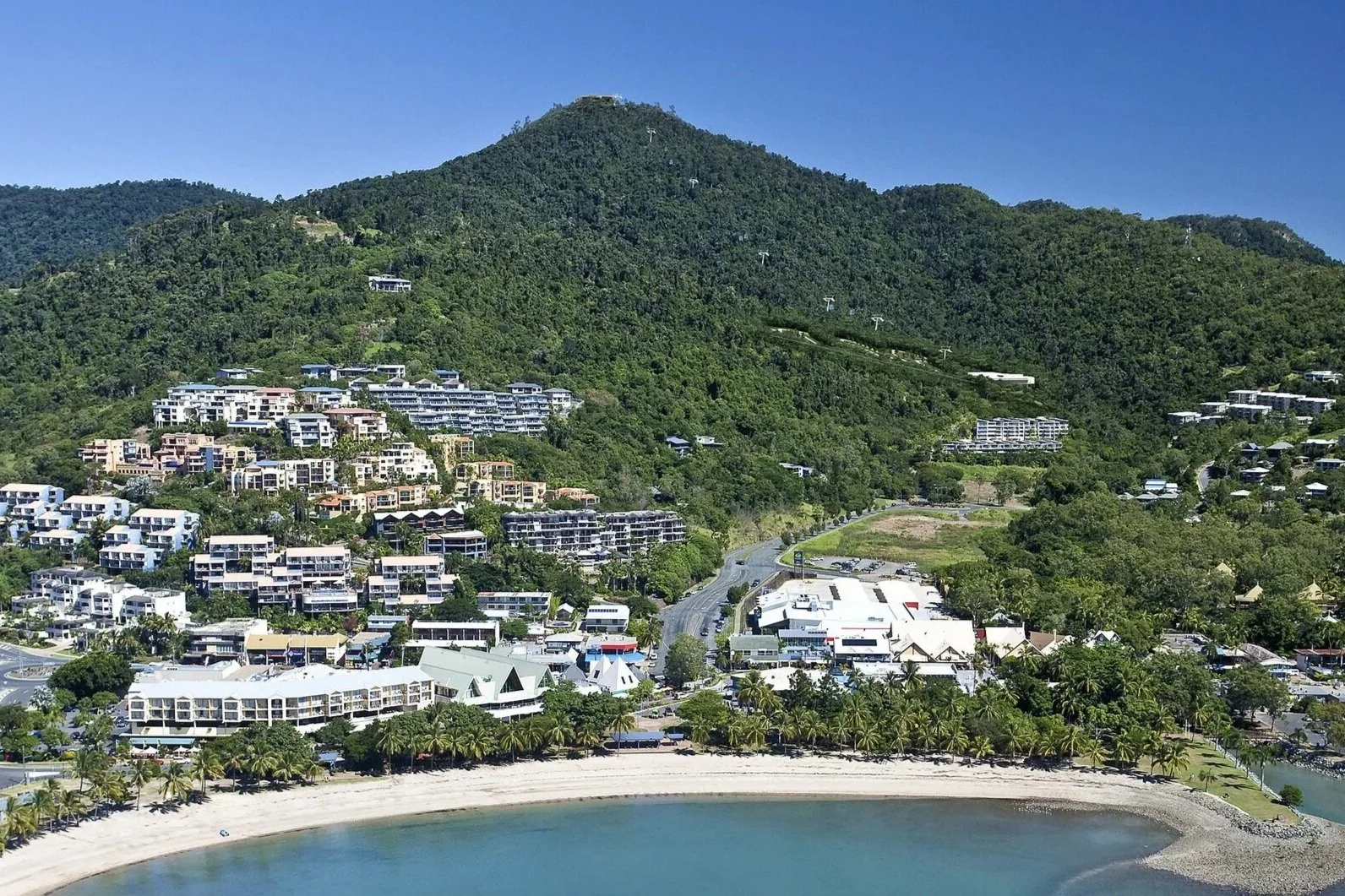 View of a coastal hillside town with multiple white and beige buildings, surrounded by green trees and a mountain in the background, beneath a clear blue sky.