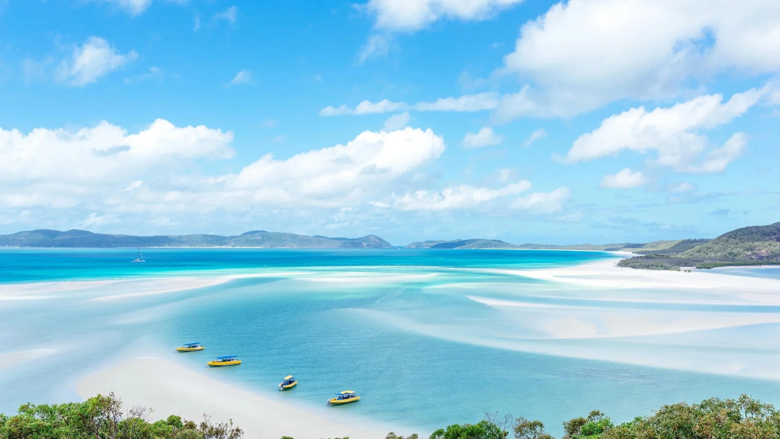 A scenic coastal view with turquoise waters, white sandy beaches, green shrubbery, distant hills, and a bright blue sky with scattered clouds.