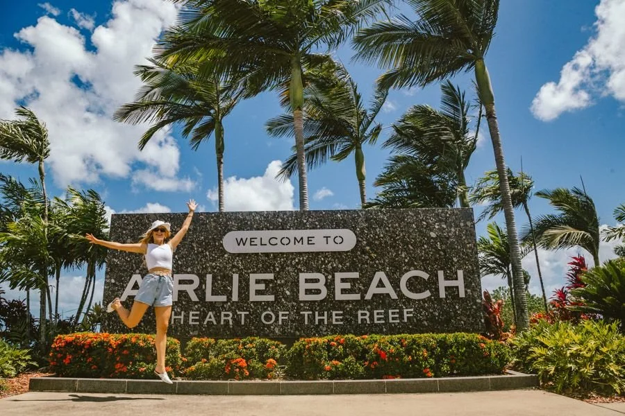 Woman in white tank top, shorts, and hat jumping next to the welcome sign at Arlie Beach surrounded by palm trees and tropical plants under a cloudy sky.