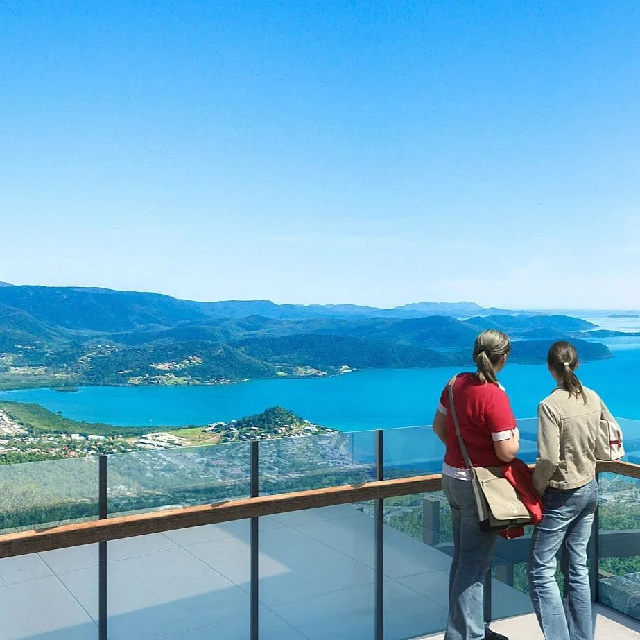 Two women stand on an observation deck overlooking a large lake surrounded by mountains and hills, with a clear blue sky overhead.