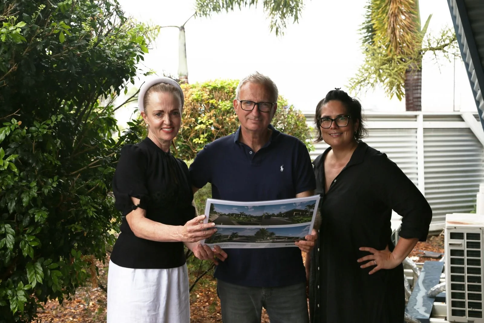 Three people standing outdoors, two women and a man, holding a large printed architectural rendering or landscape plan, with greenery and a building in the background.