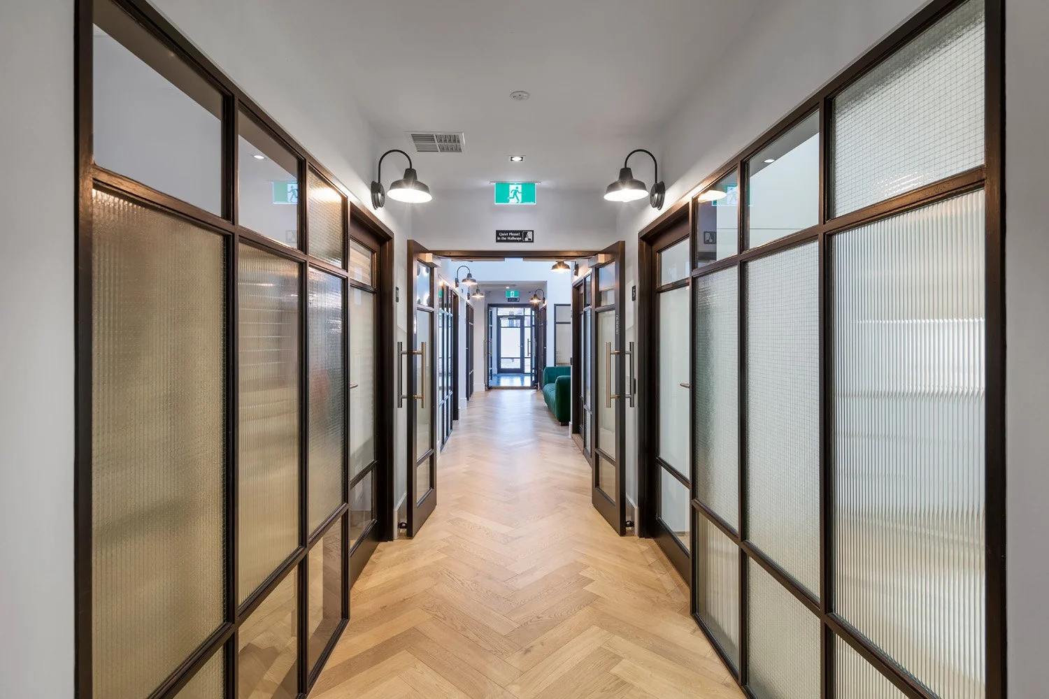 Empty corridor in a modern office or hotel with glass partition walls, wooden floors, wall-mounted lamps, and exit signs at the end.